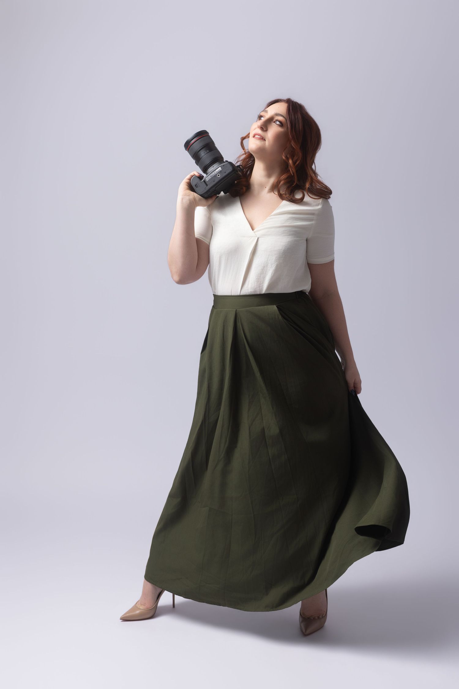 Woman in white top and olive maxi skirt holding a DSLR camera, posing gracefully on a light gray background.
