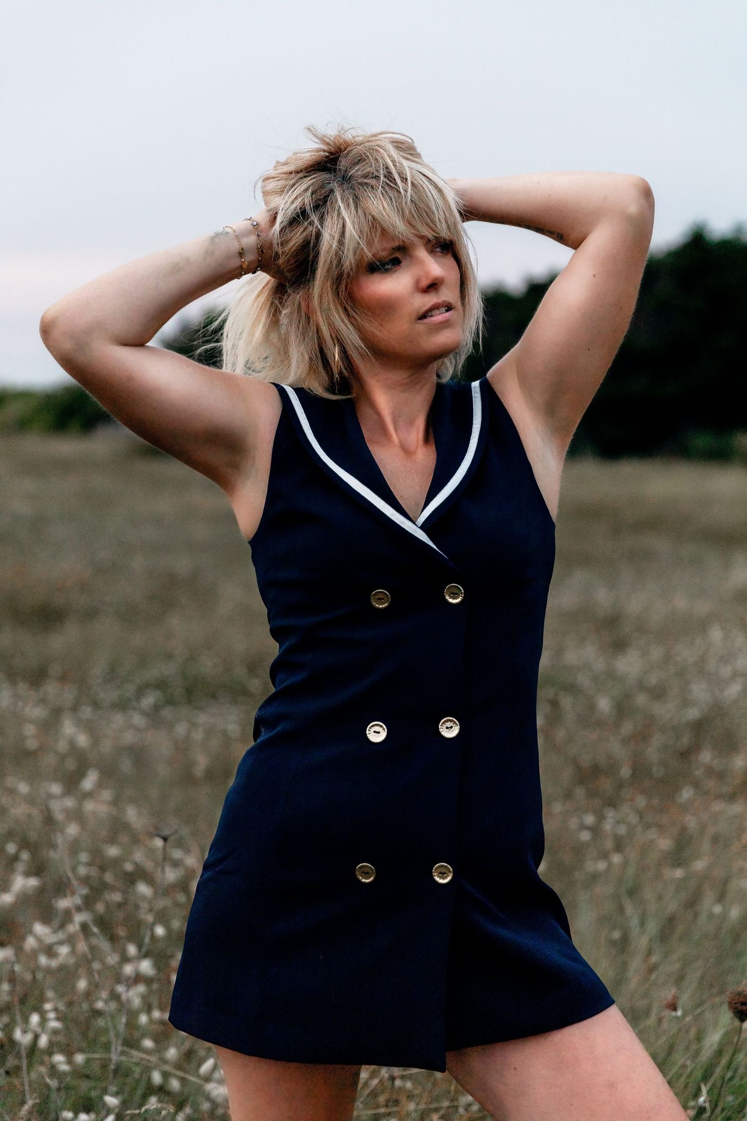 Stylish person in a navy blue sleeveless dress poses playfully in a grassy field on a bright summer day.