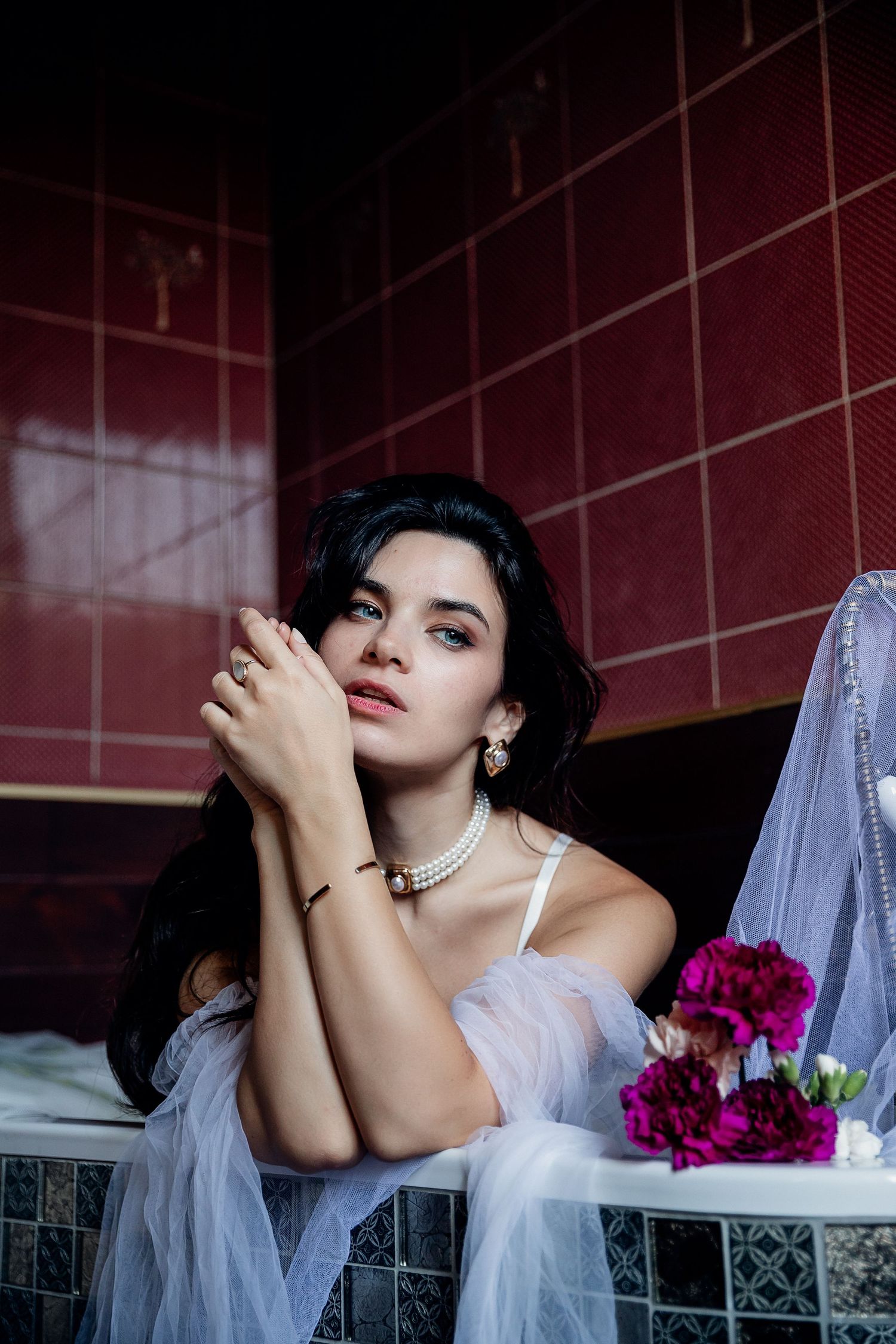 Artistic bridal photoshoot in dramatic lighting against red tiles features white veil and vibrant purple flowers.