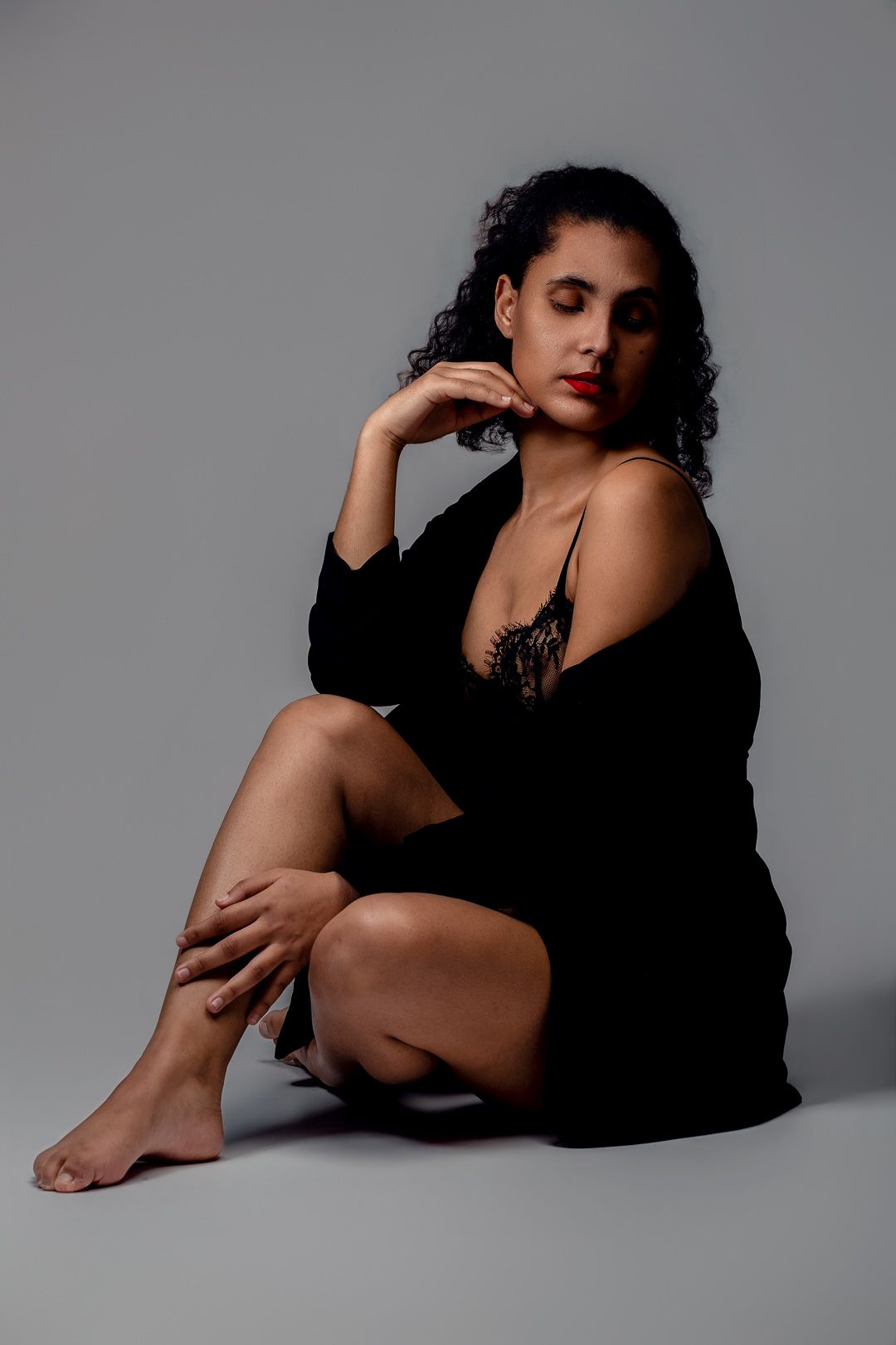 Woman in black lingerie and robe sitting gracefully on studio floor with curly hair and red lips.