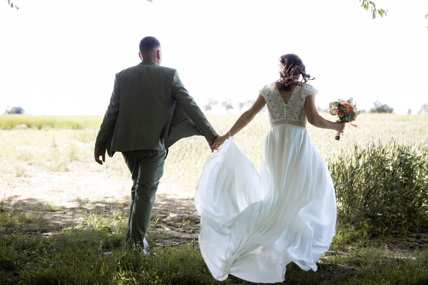 A bride in a flowing white gown and groom in a green suit walk hand-in-hand through a scenic outdoor field.