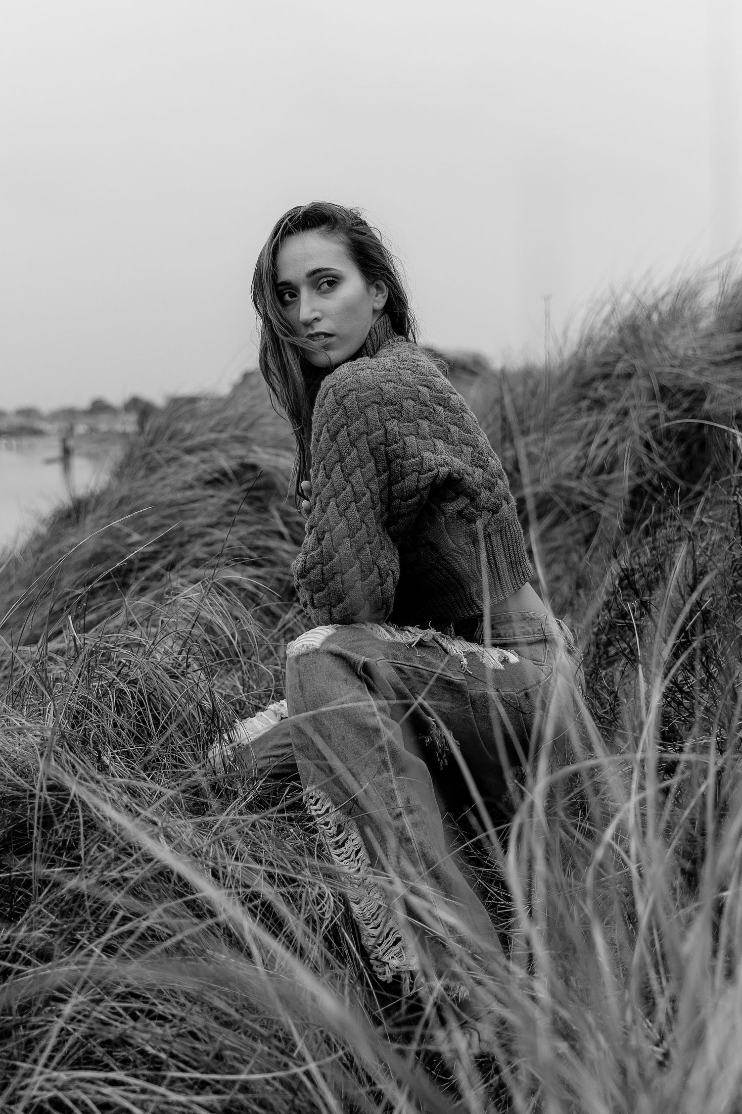 Black and white portrait photograph of person sitting in coastal beach grass wearing a sweater.