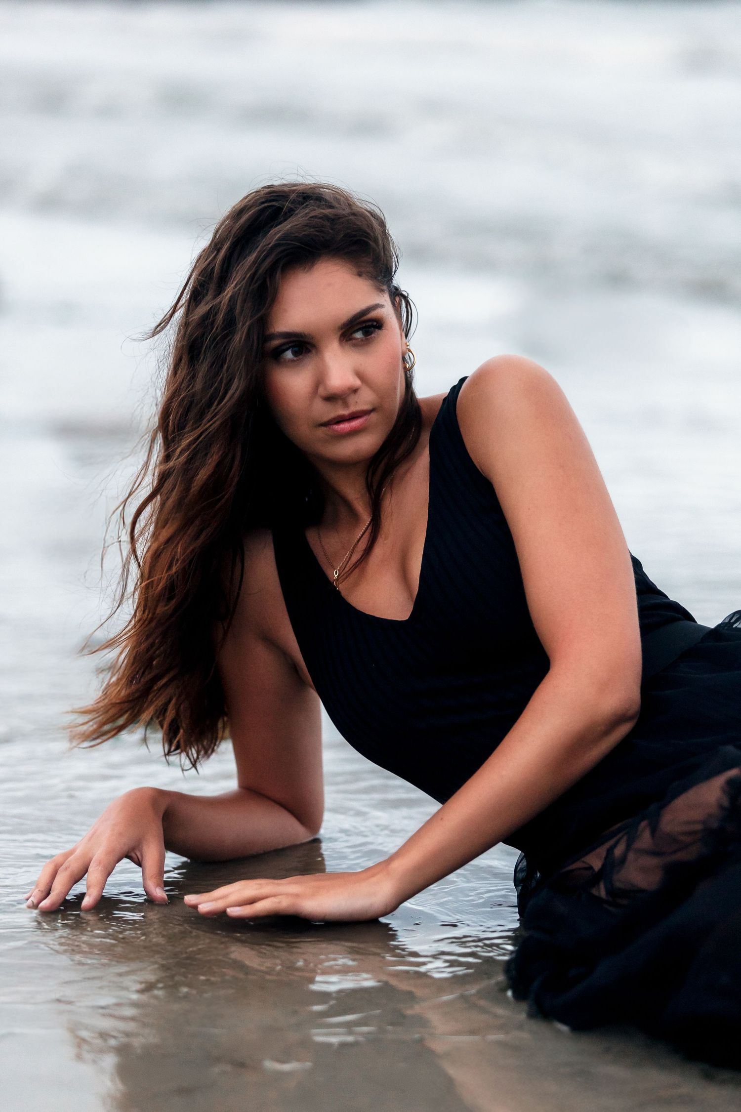 Woman in black swimsuit poses laying on wet sand at the beach during overcast day.
