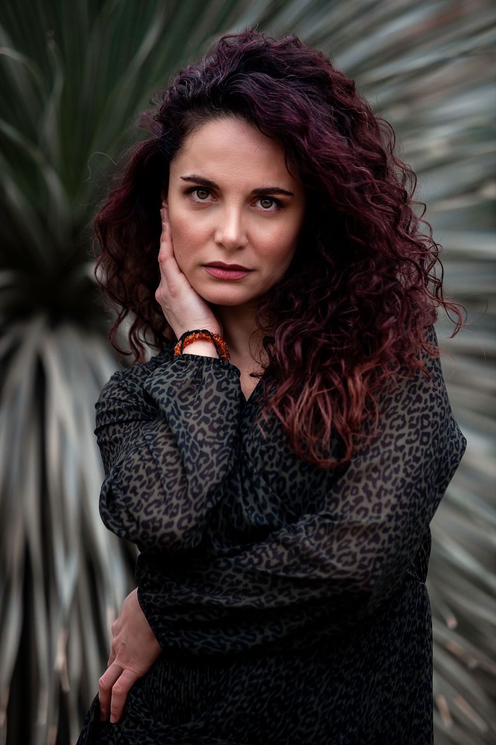 Woman with curly red-purple hair in leopard print blouse poses thoughtfully against tropical plant backdrop.