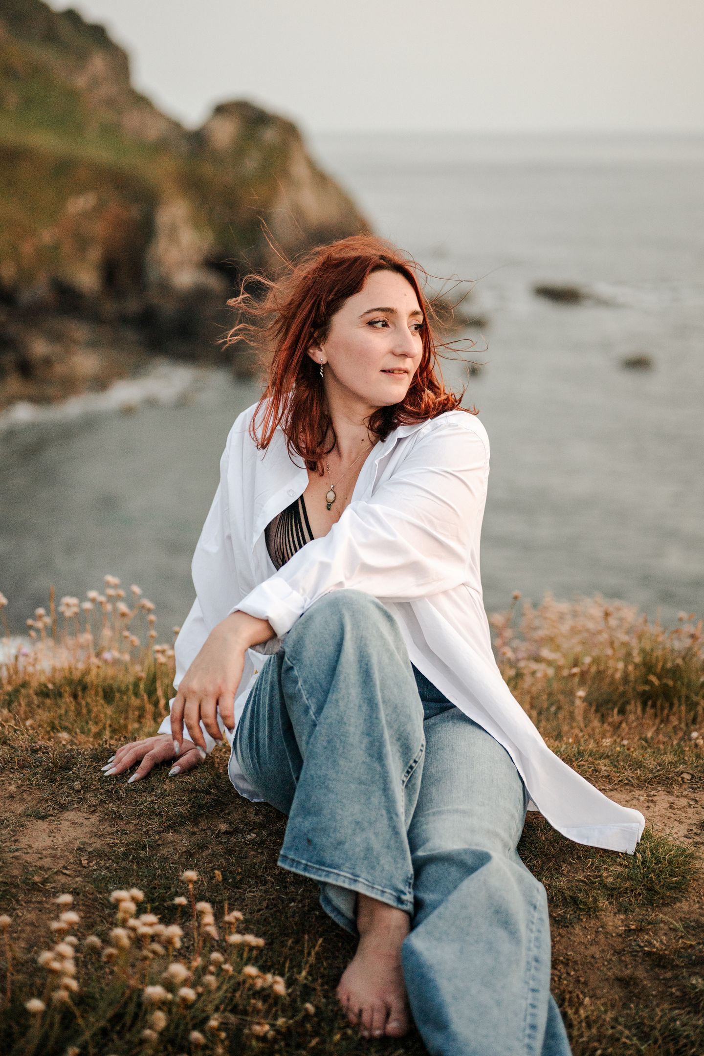 A person in blue jeans and white sweater sits on a coastal cliff edge during golden hour at sunset.