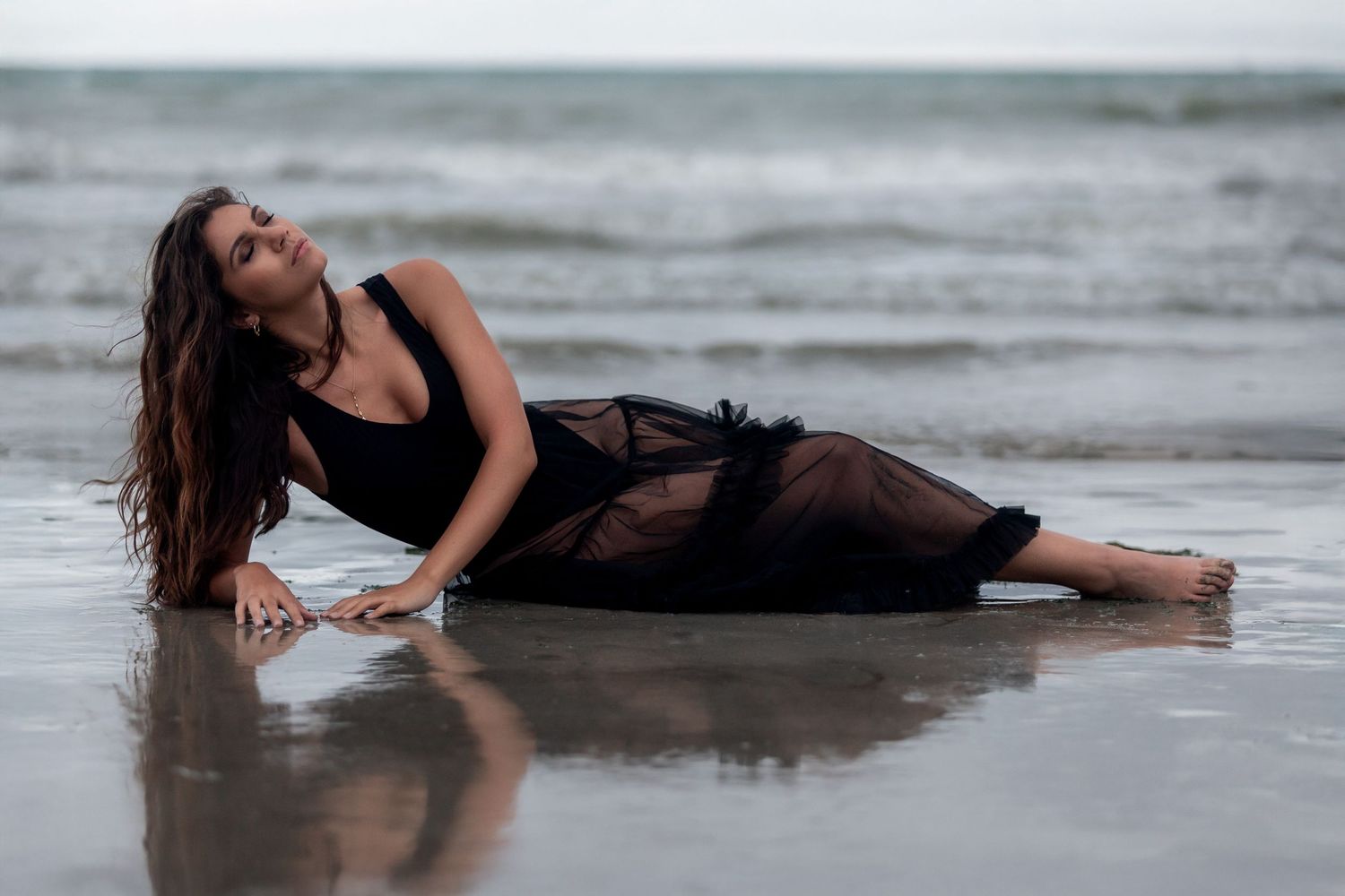 A woman in a black sheer dress lies gracefully on wet sand at the beach during overcast conditions.