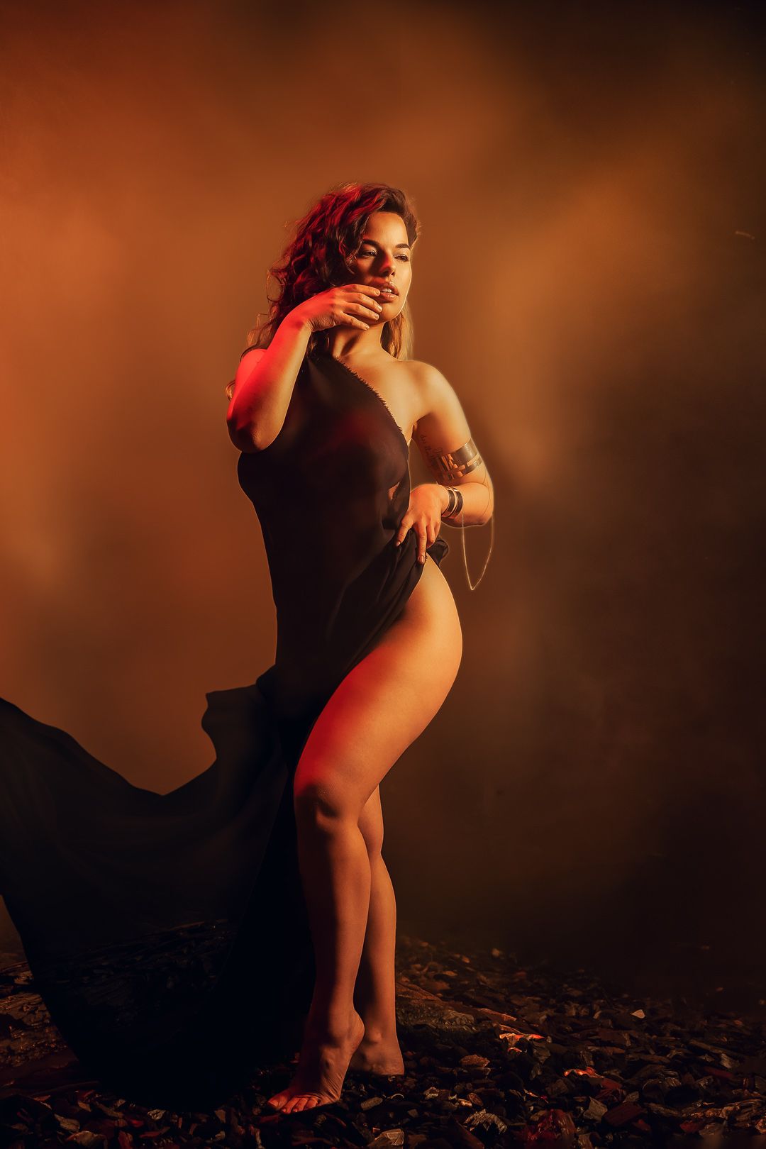 A woman in a flowing black dress poses dramatically against a smoky orange-red backdrop with moody atmospheric lighting.