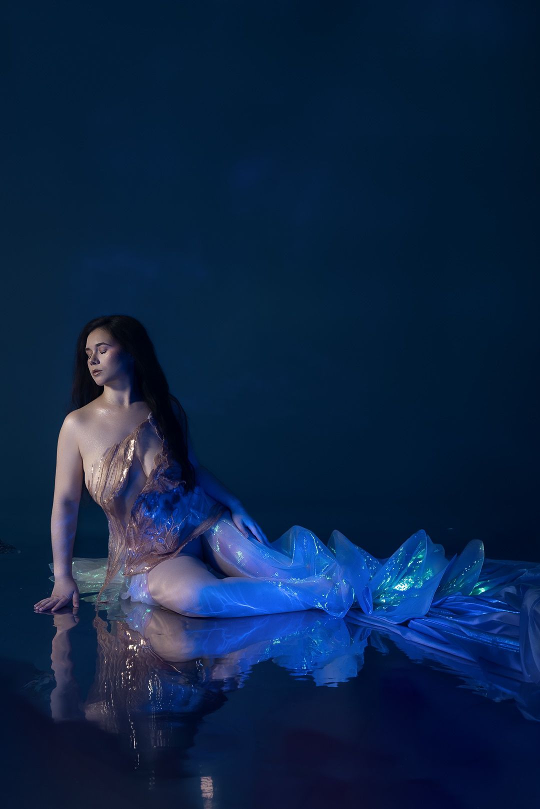 A woman in a glowing blue mermaid gown poses gracefully on a reflective surface against a dark blue background.