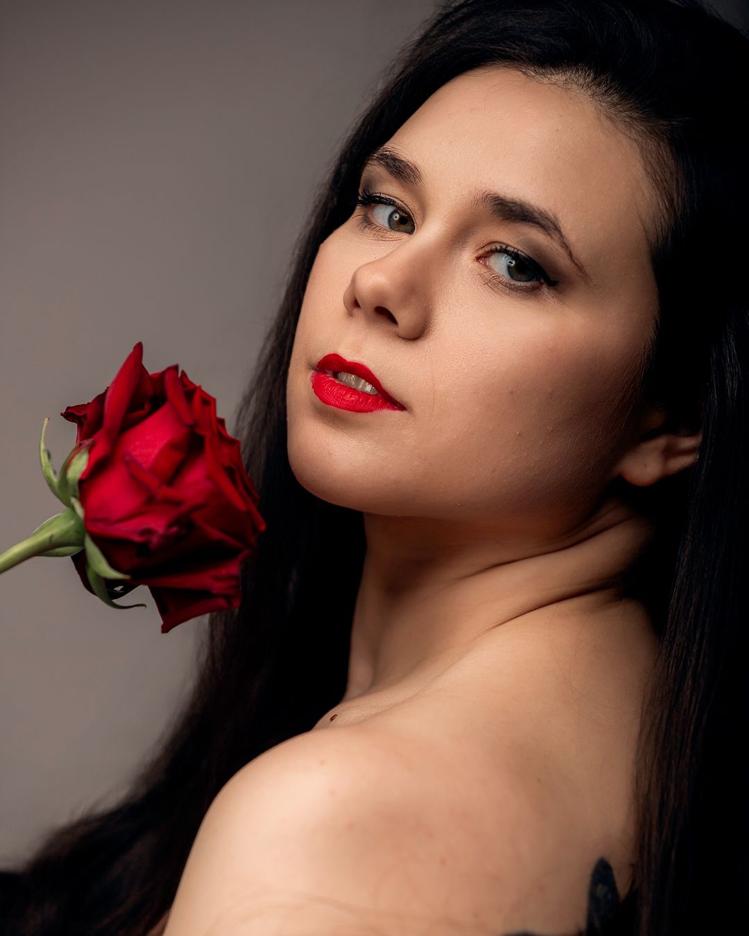 A woman with red lips and dark hair holds a red rose, gazing softly at the camera against a gray background.