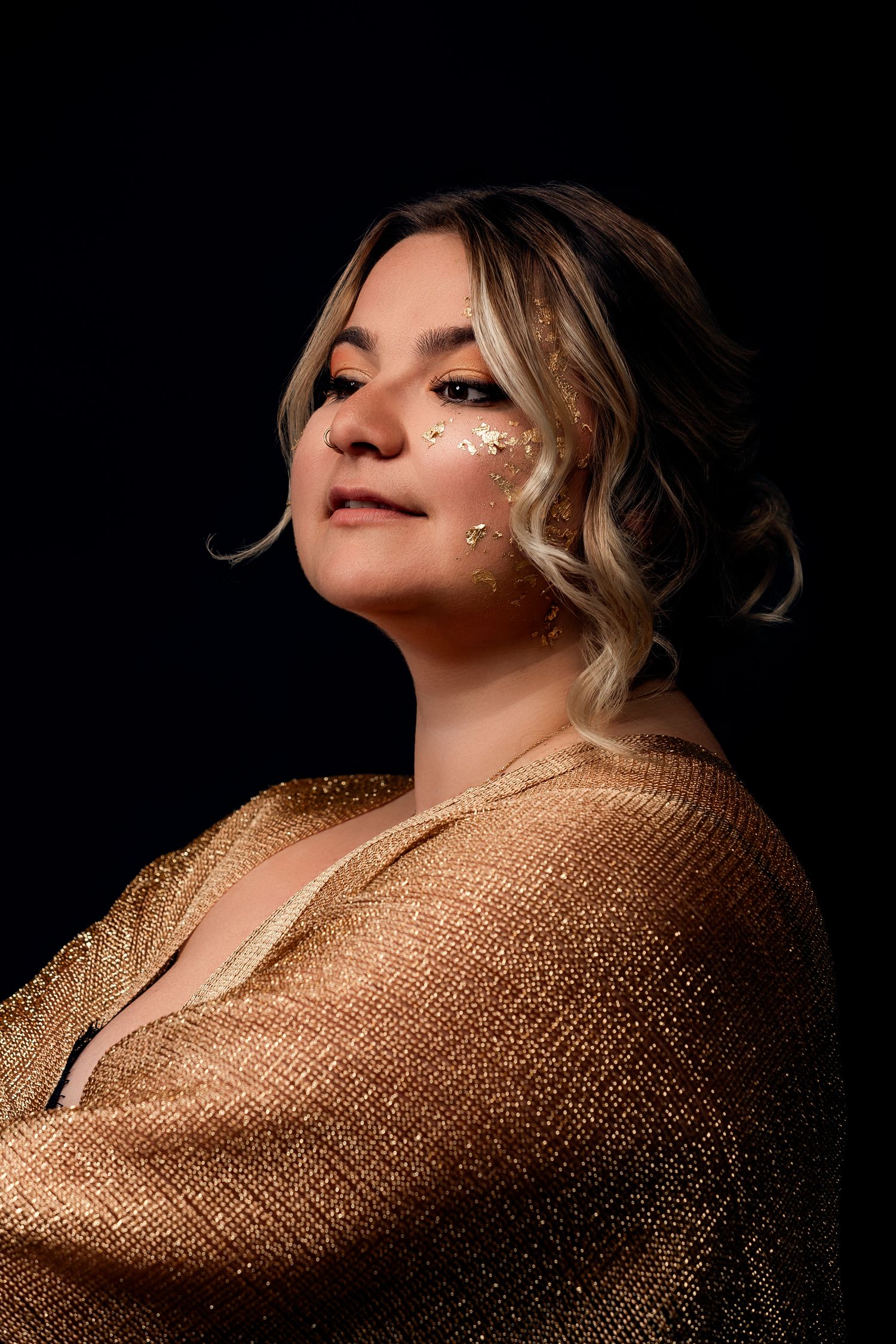 Woman in gold shimmer wrap with glitter face makeup poses confidently against dark background.