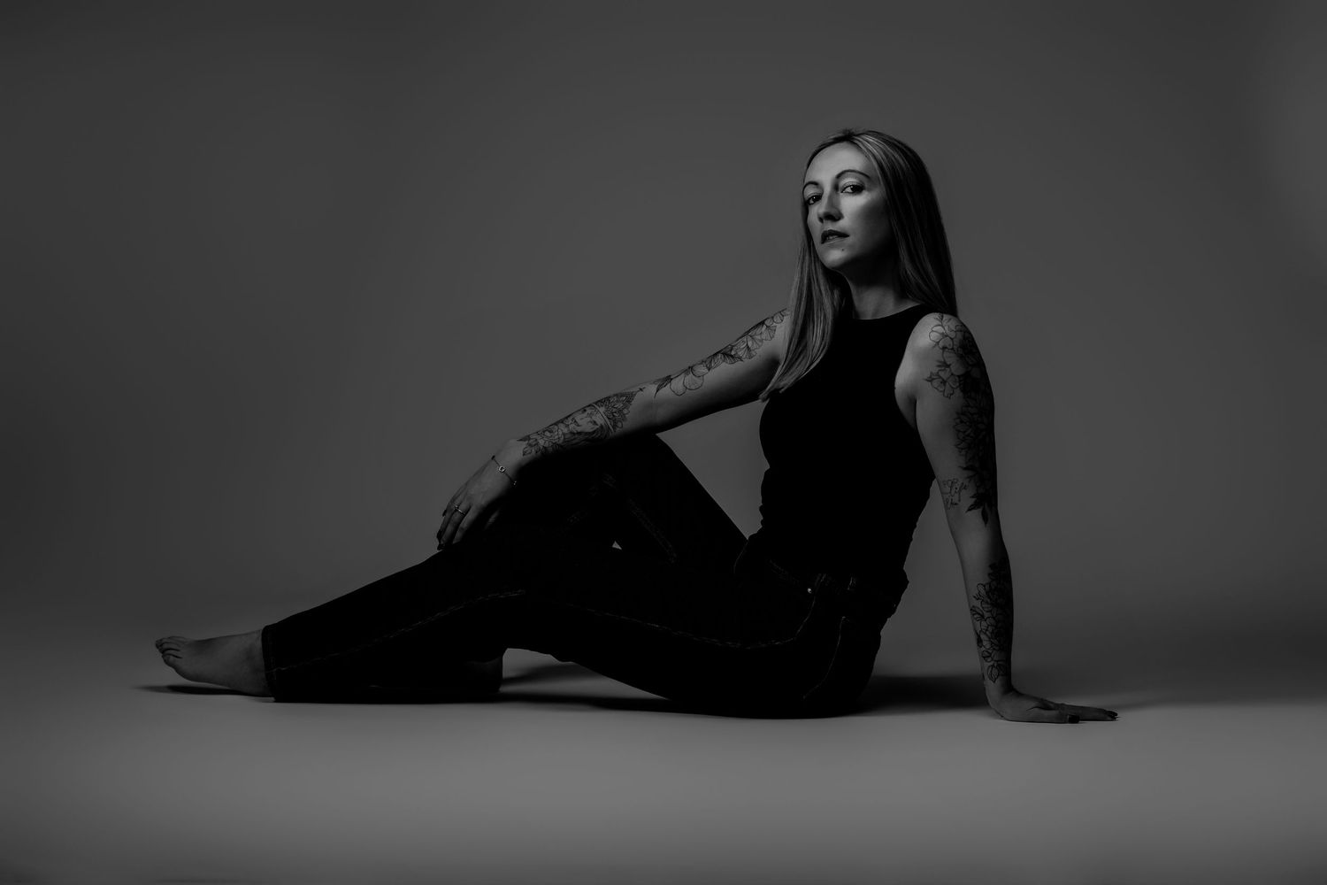 Black and white studio portrait of a tattooed woman in black outfit sitting gracefully on the floor.