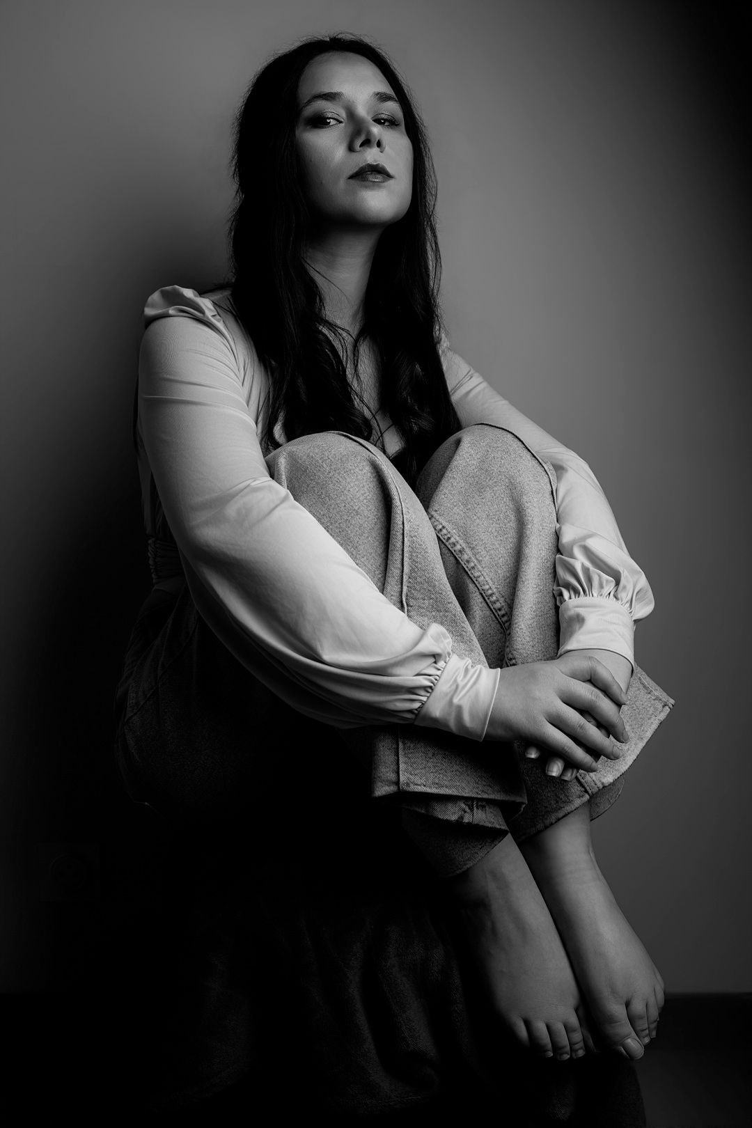 Black and white portrait of a young woman with long dark hair sitting against a wall hugging her knees.