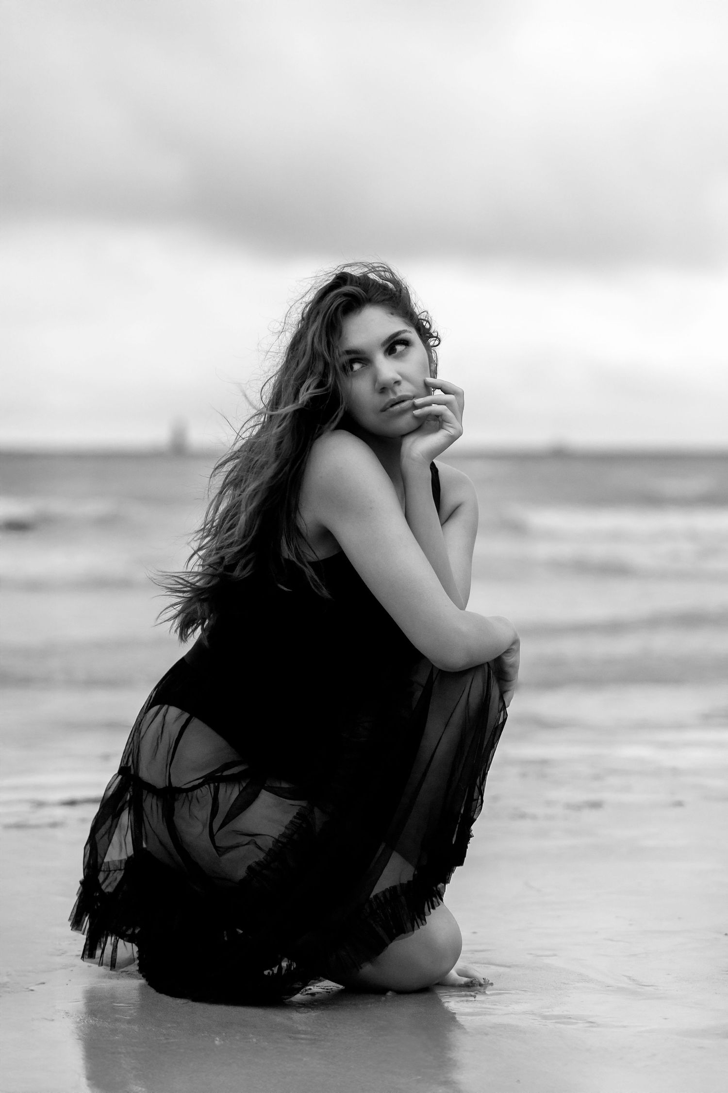 A silhouetted figure in a black dress poses dramatically on a beach in black and white photography.