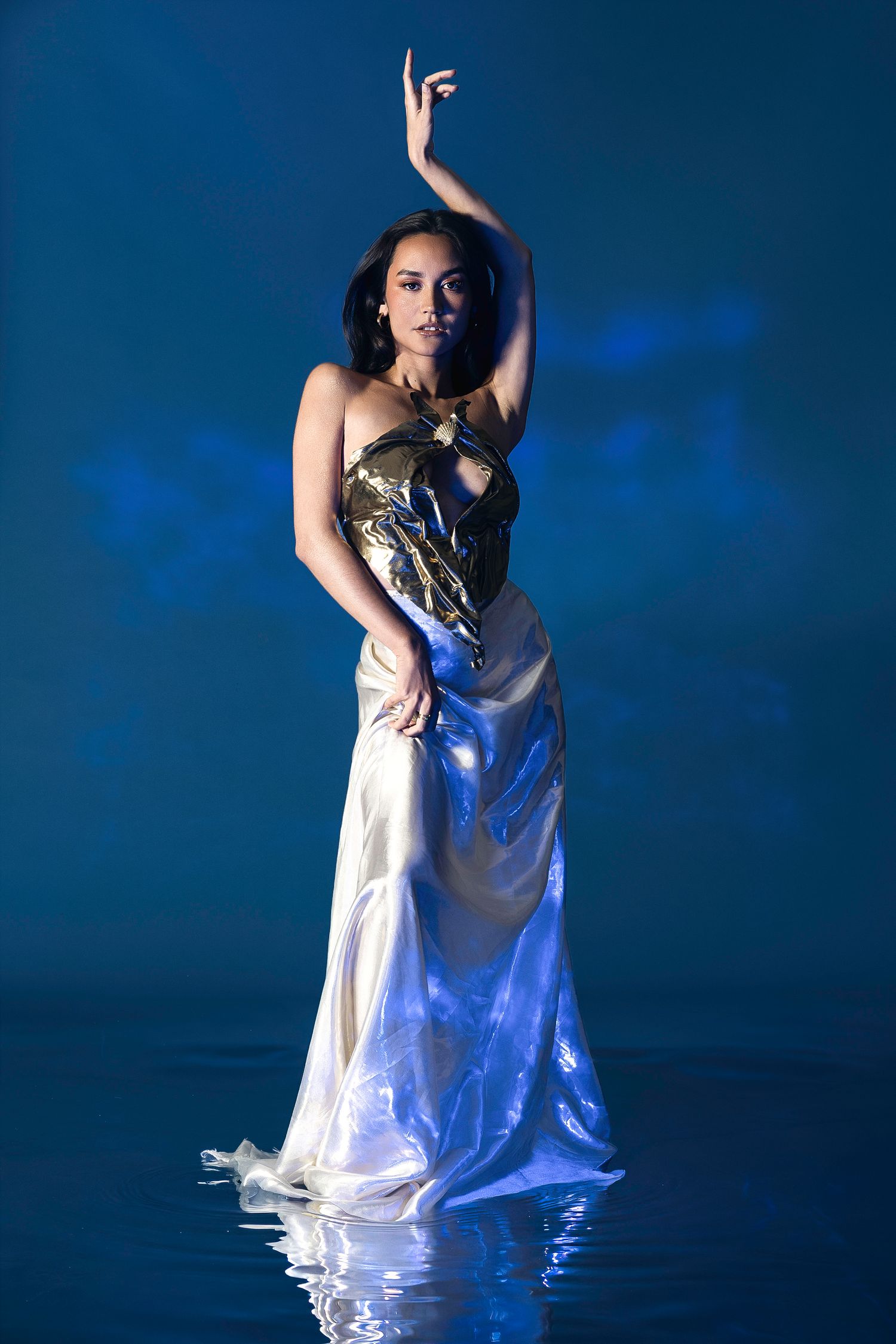 Woman in gold and white gown poses gracefully against a blue backdrop with water reflections.