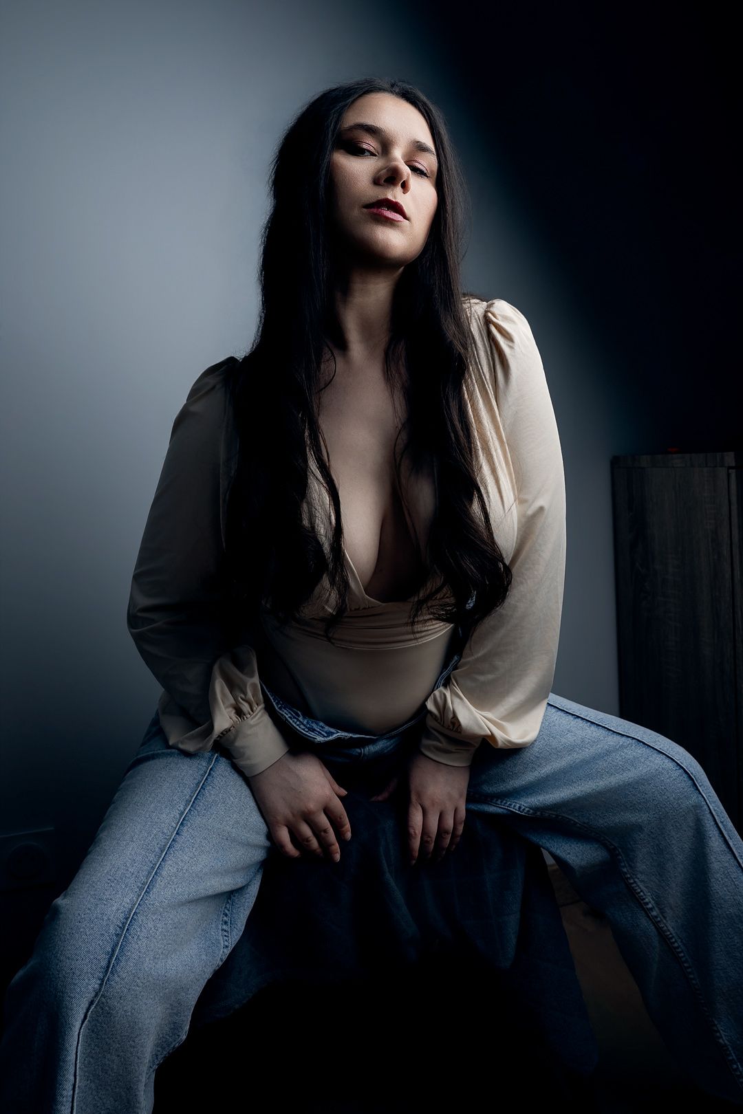 Young woman with long dark hair poses seated in dramatic moody lighting, wearing cream blouse and wide-leg jeans.