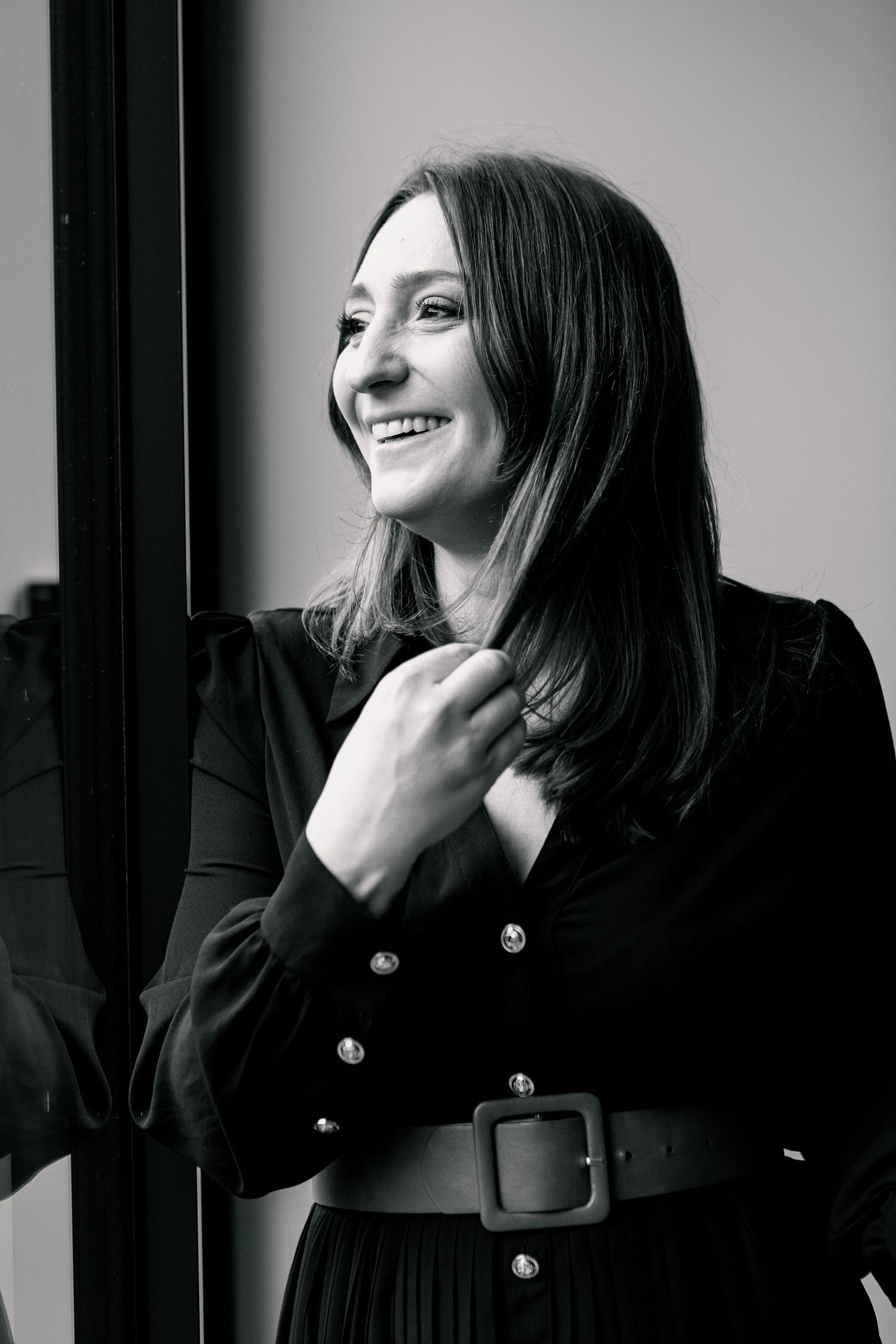 A black and white portrait of a woman in a belted coat smiling by a window.