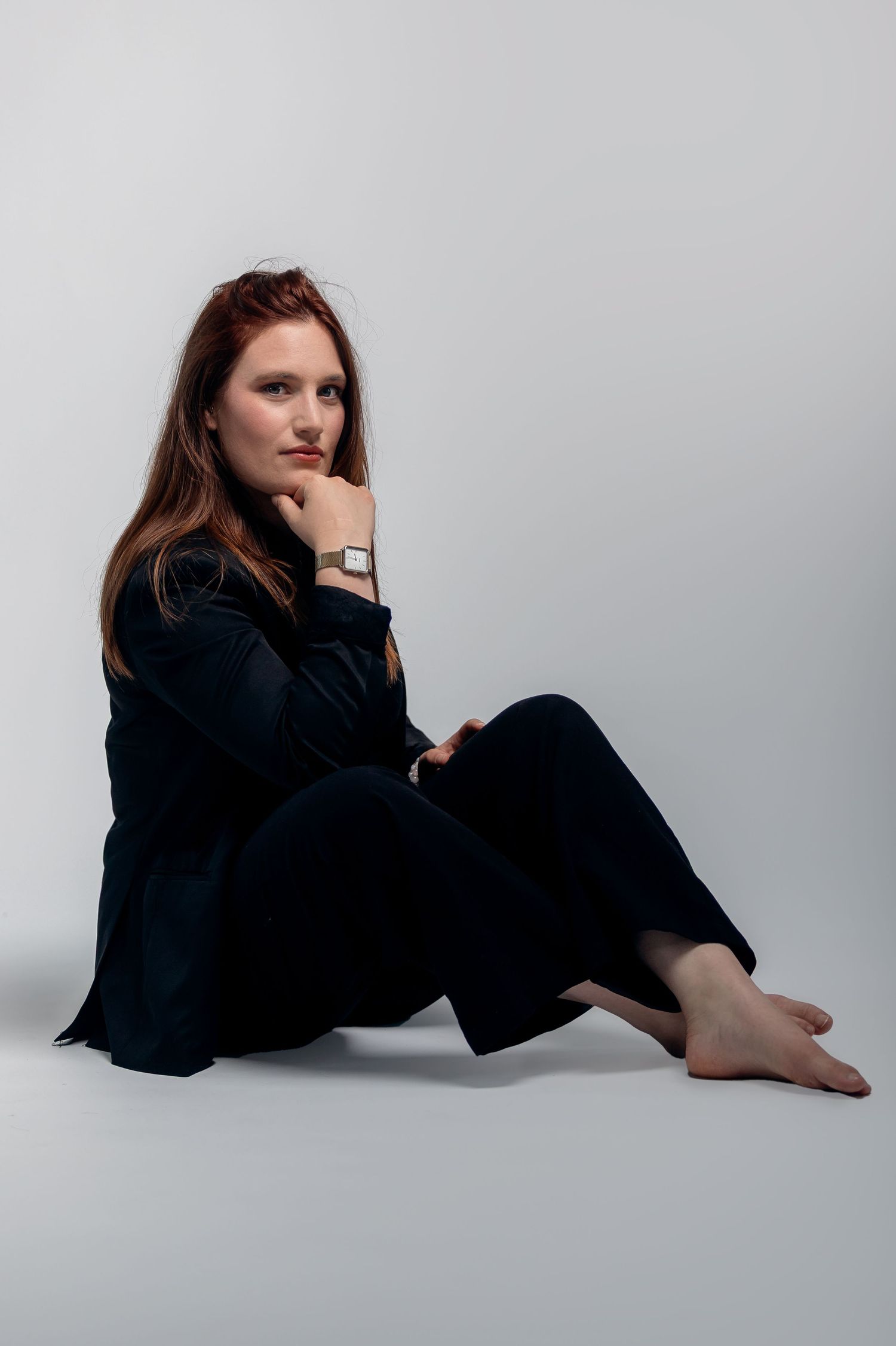 Minimalist portrait against light gray background showing person in black outfit sitting on floor with bare feet.