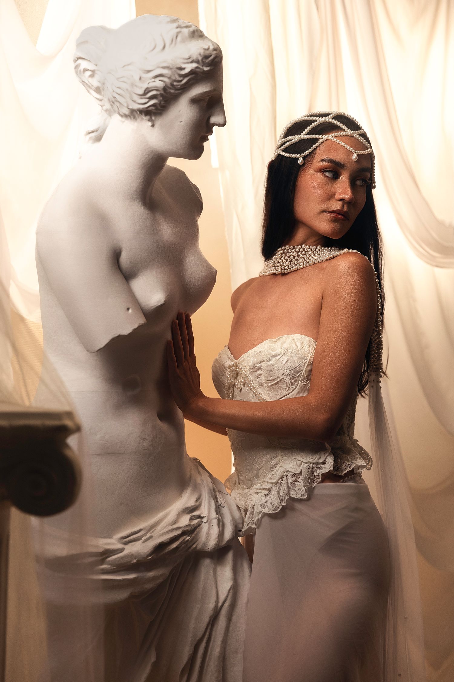 Woman in white corset dress poses beside Venus de Milo statue, wearing pearl headpiece and choker necklace.