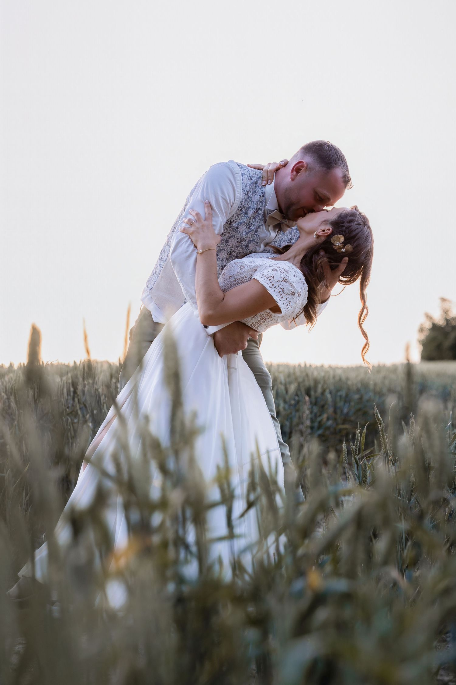 Bride and groom sharing a romantic dip kiss in a golden wheat field during sunset on their wedding day.