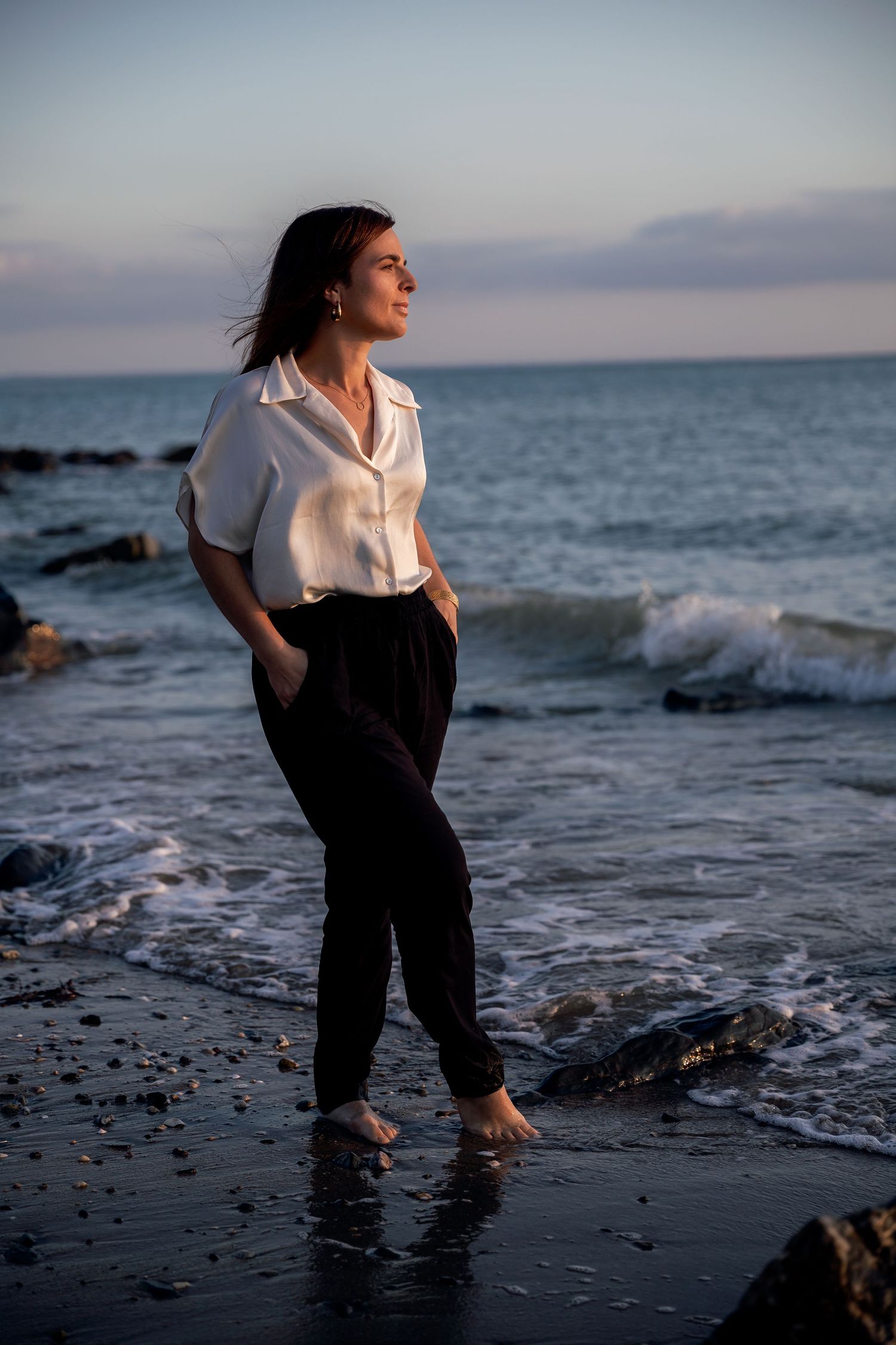 Woman in white shirt and black pants stands barefoot on rocky beach at sunset, gazing at ocean waves.