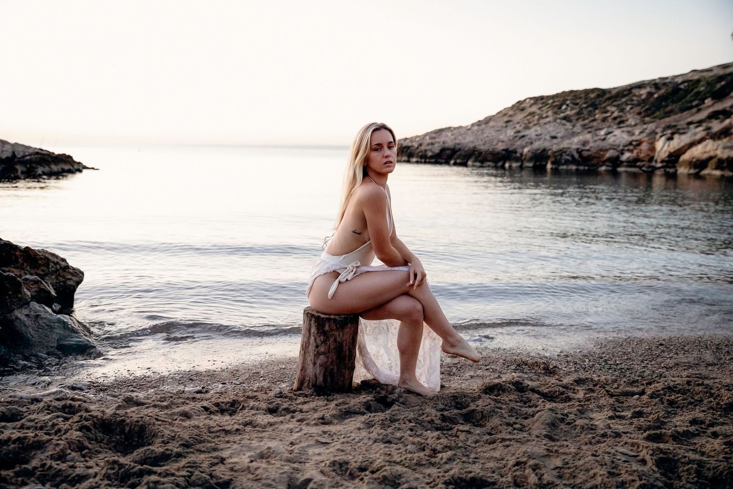 A blonde woman in a white bikini sits on a tree stump on a sandy beach at sunset.