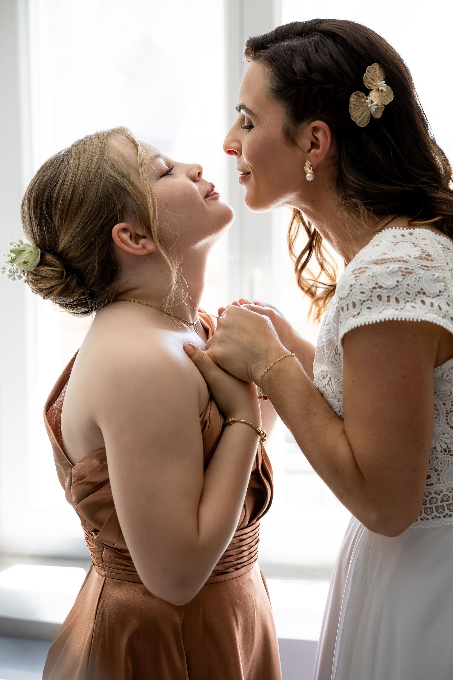Bride in white lace dress holds hands with bridesmaid in bronze gown, sharing a tender moment near a bright window.