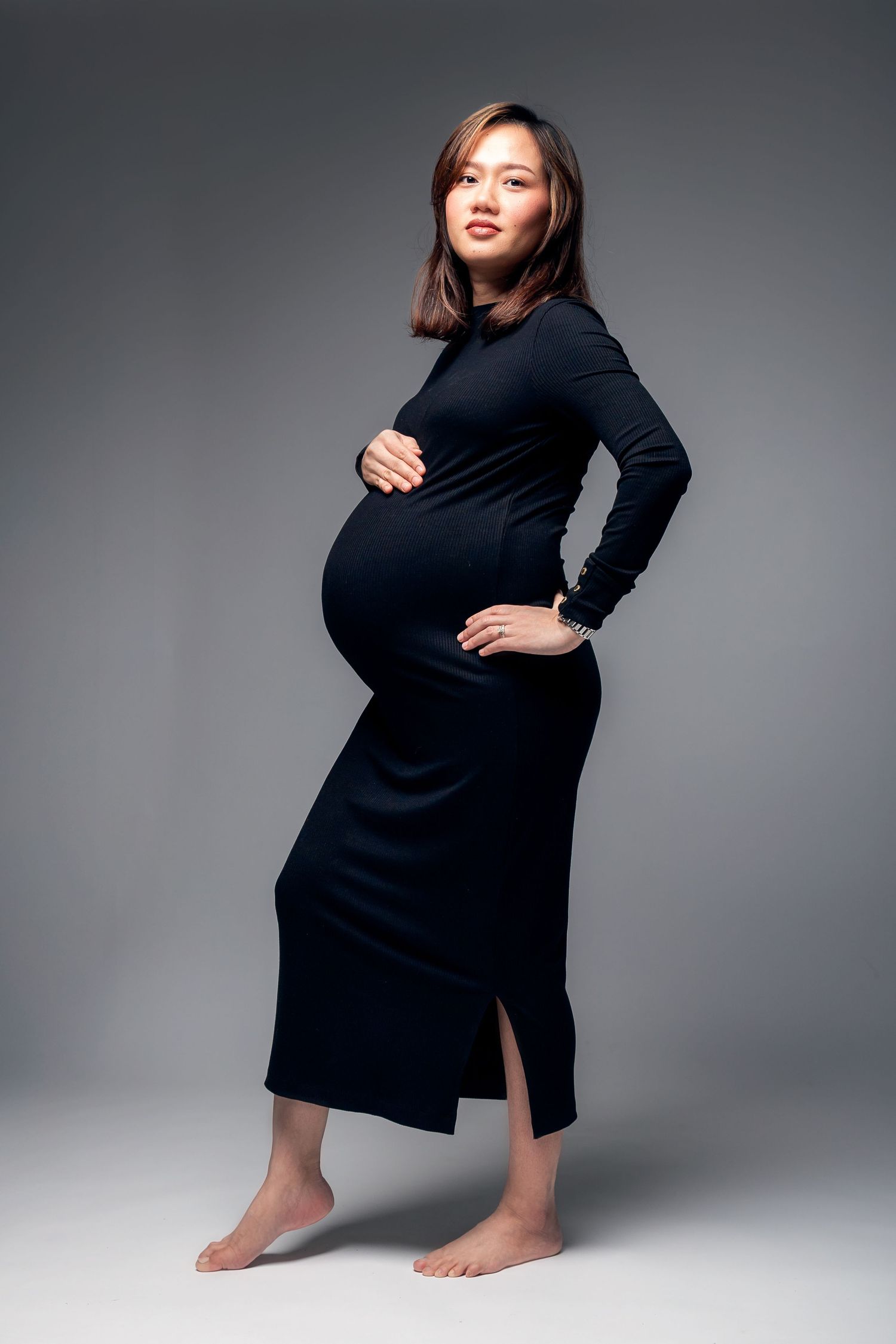 Pregnant Asian woman in black long-sleeve dress cradling baby bump, standing barefoot against gray studio background.