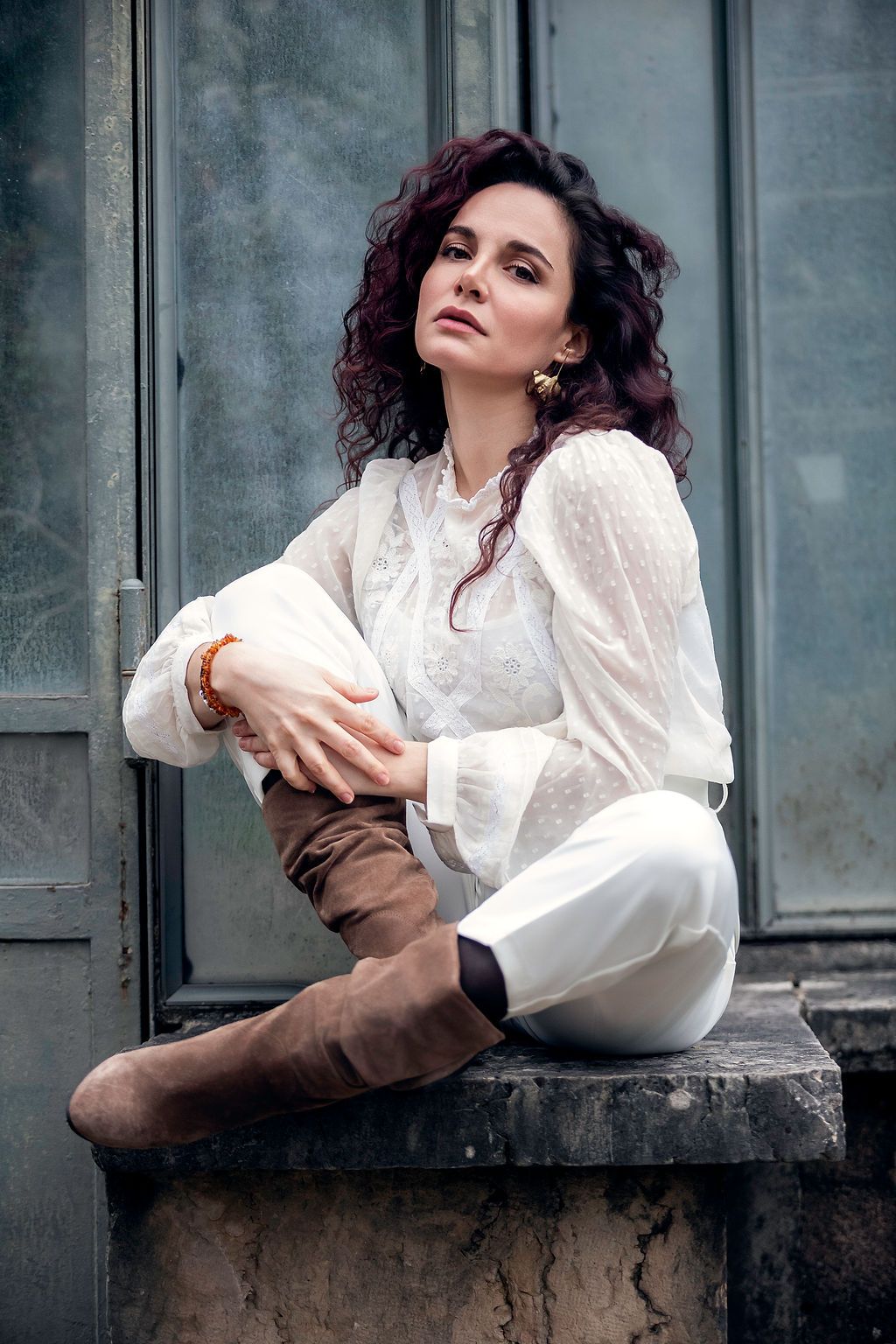 A woman with curly auburn hair sits on a stone ledge wearing a white blouse, white pants, and brown knee-high boots.