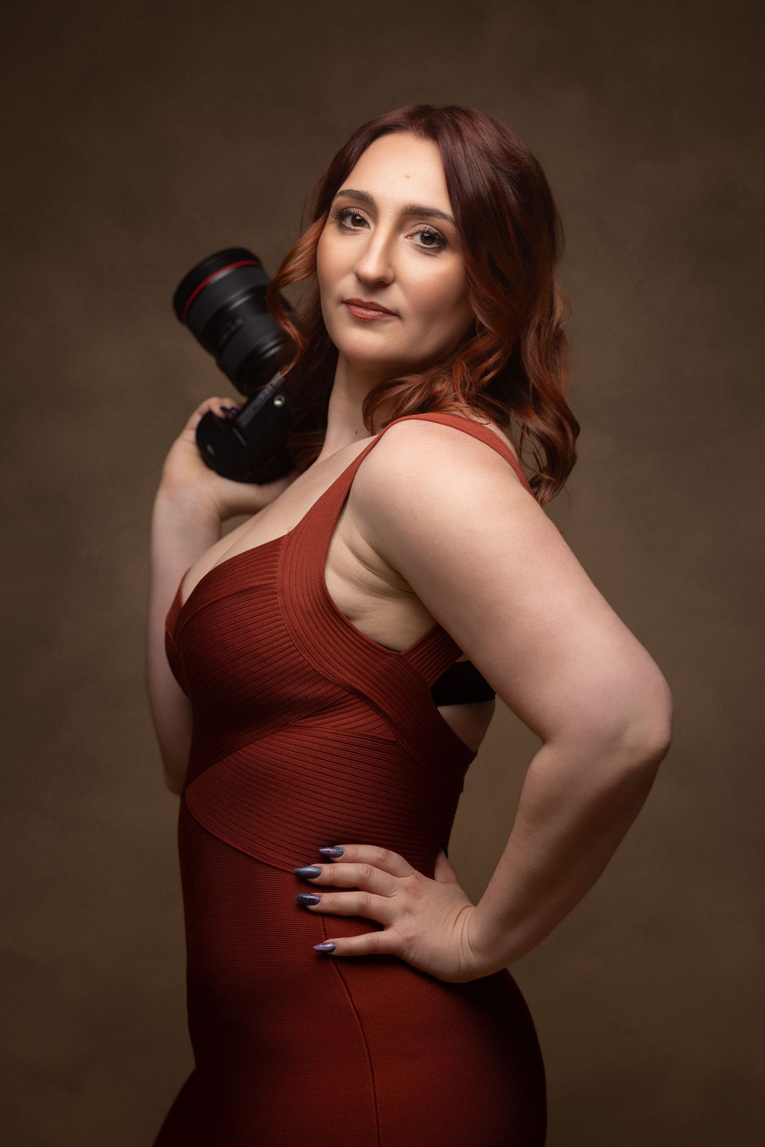 A woman in a red dress holds a camera over her shoulder, posing confidently in a studio portrait.