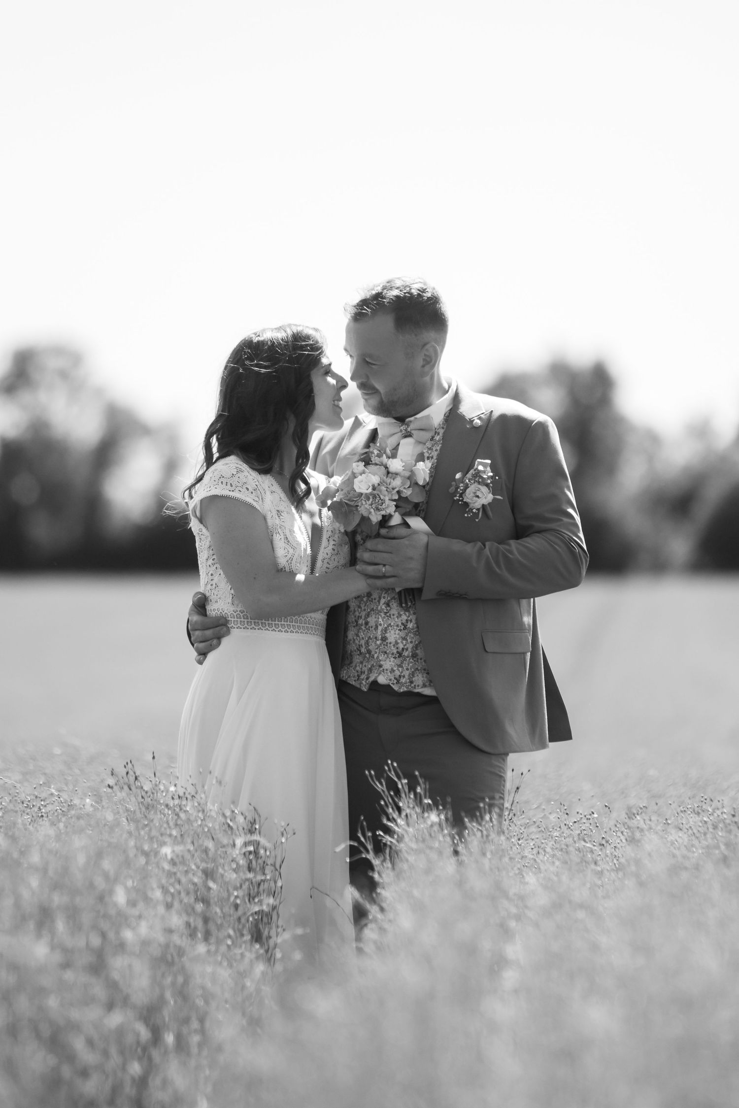 Black and white photo of a bride and groom embracing in a field, holding a bouquet on their wedding day.