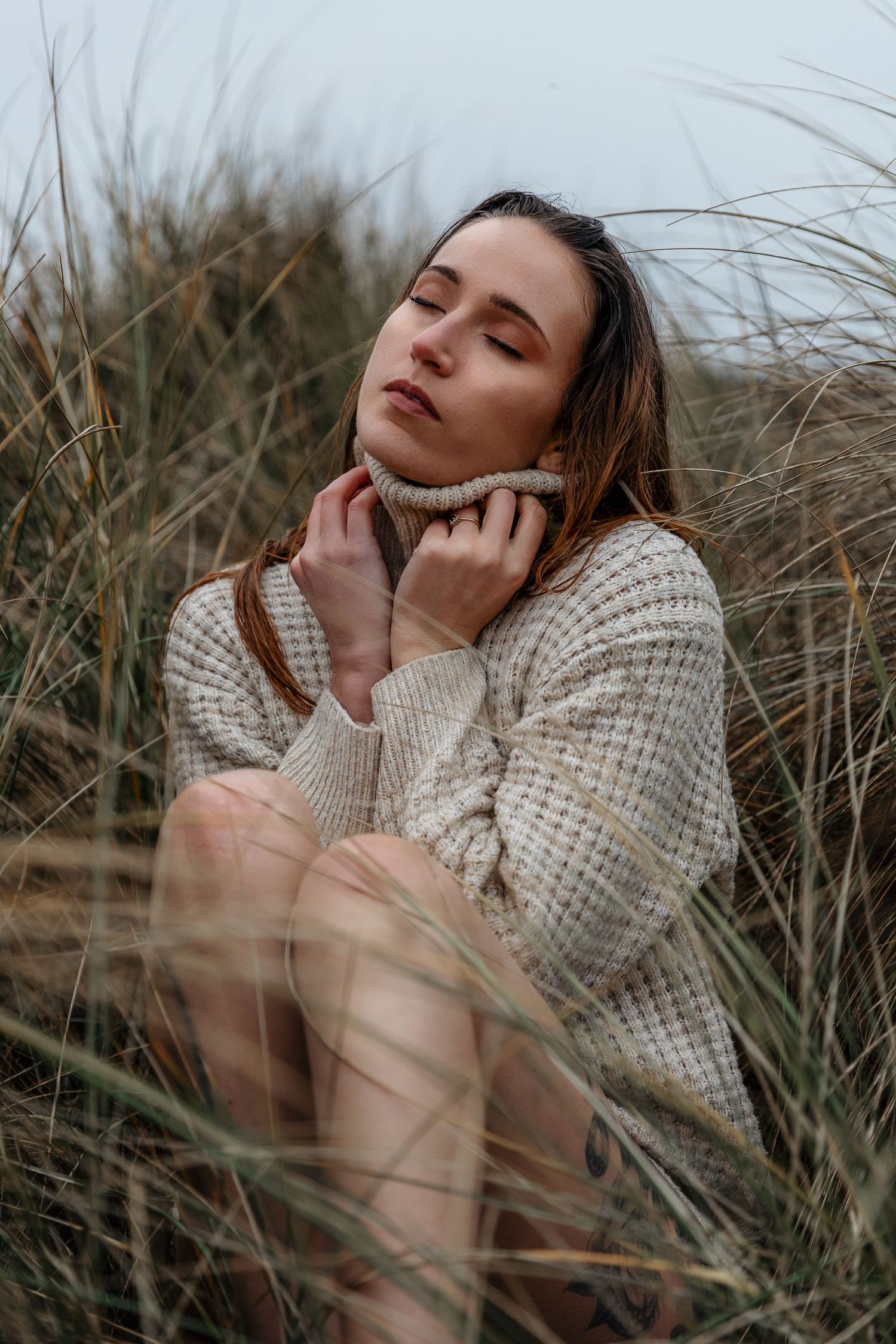 Cozy winter portrait of someone wearing a cream knit sweater sitting among tall grass at dusk.