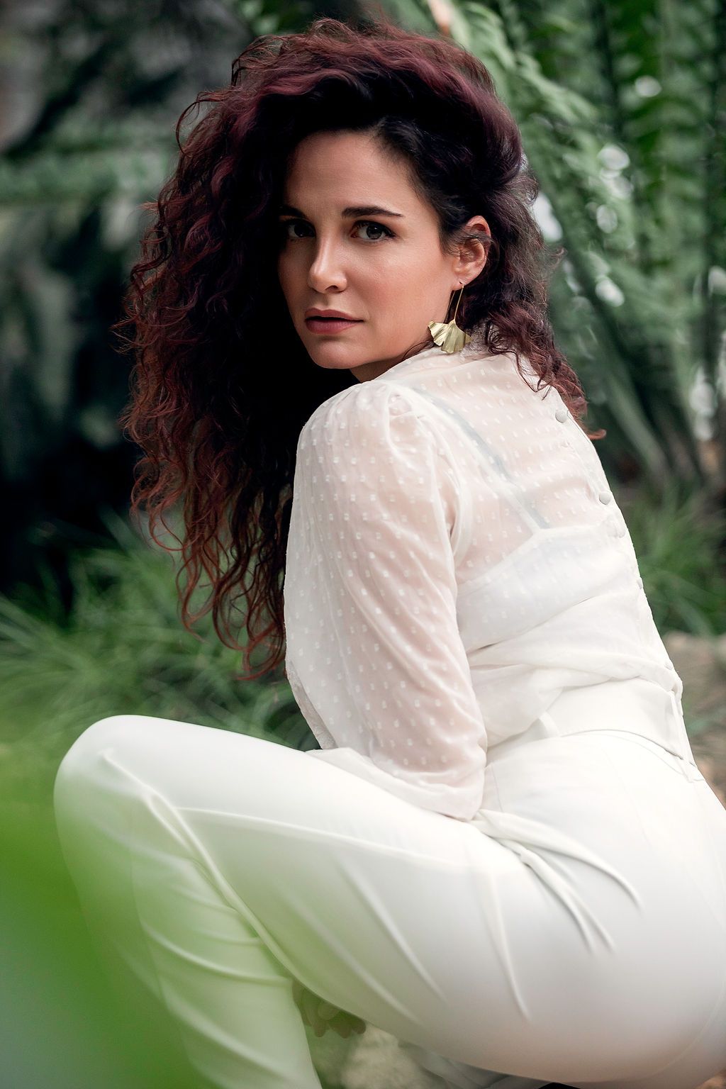 Woman with curly auburn hair in white lace blouse and pants poses outdoors among lush green foliage.