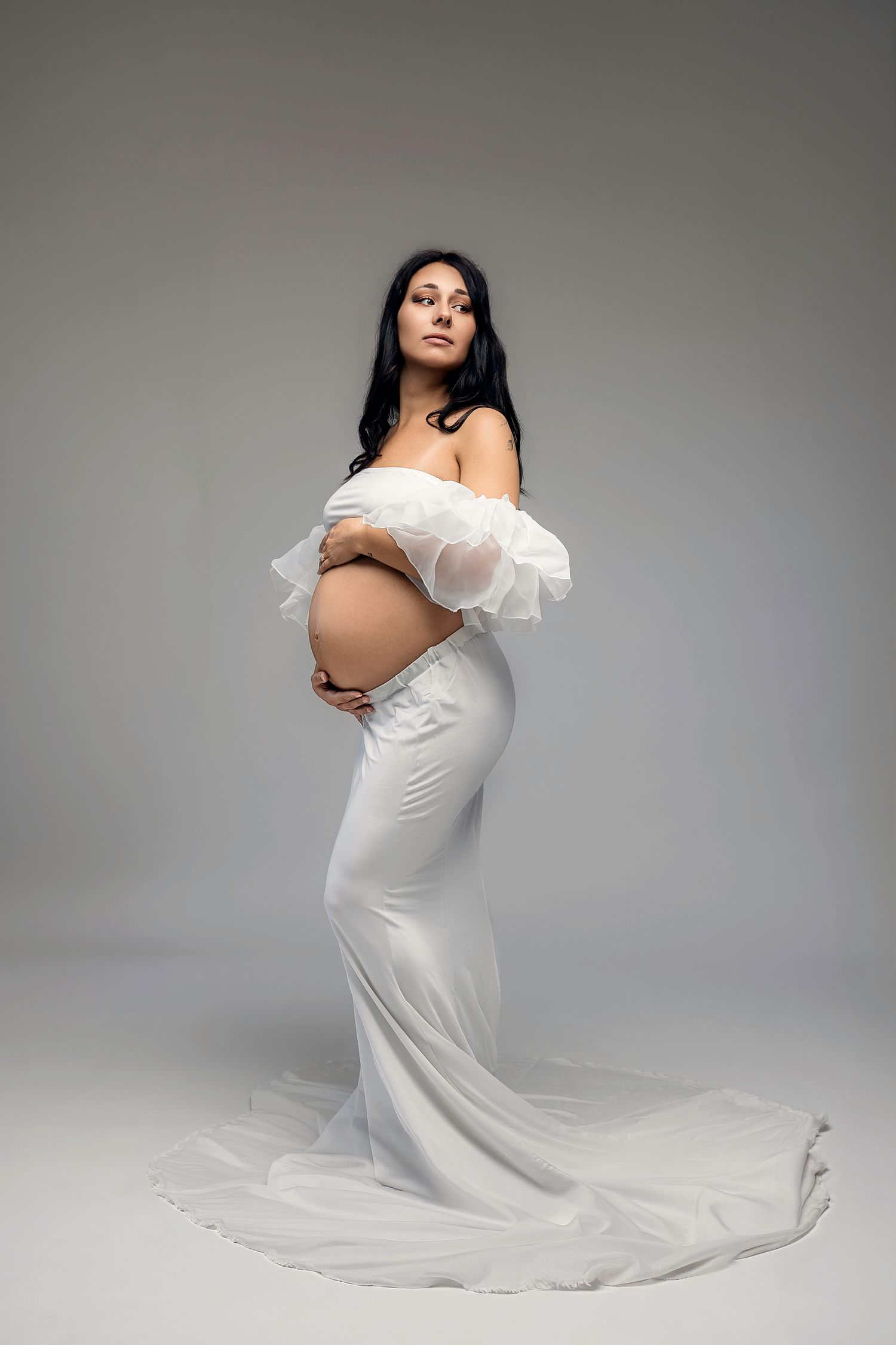 Pregnant woman in elegant white off-shoulder gown with ruffles, cradling baby bump in studio maternity photo.