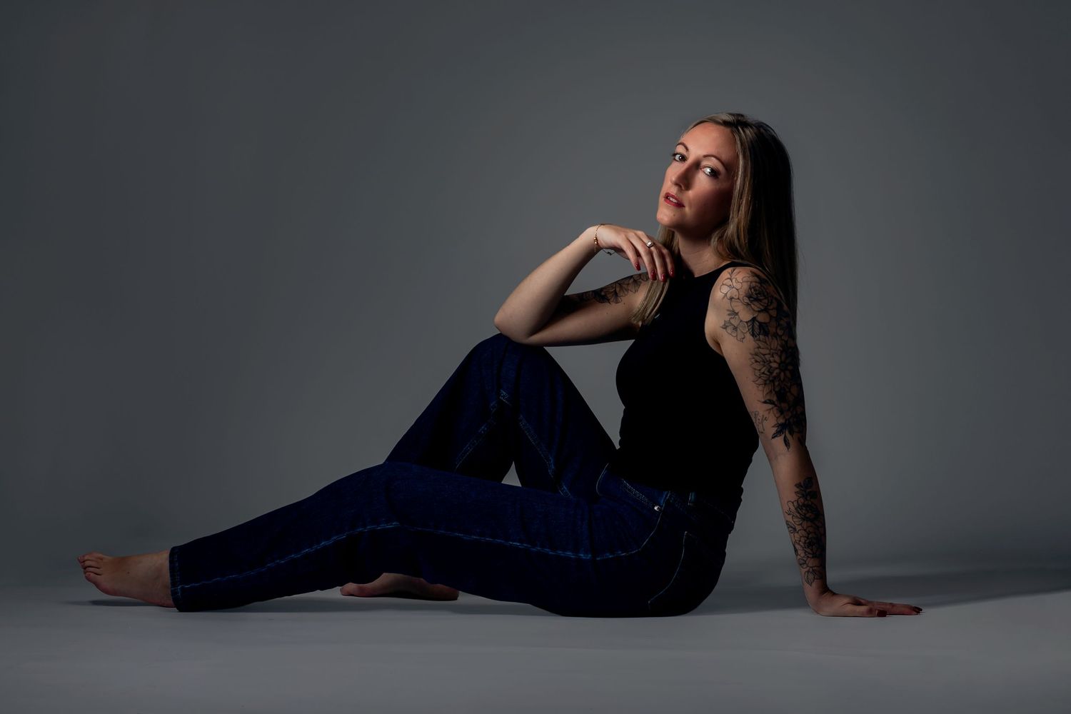 Tattooed woman in black top and blue jeans sitting gracefully on studio floor with long hair draped over shoulder.