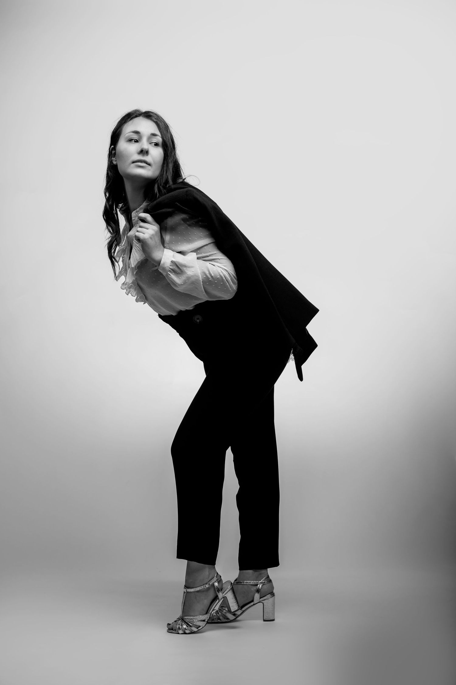 Black and white photo of a woman in dark trousers, white blouse, and blazer draped over shoulder, posing in heeled sandals.