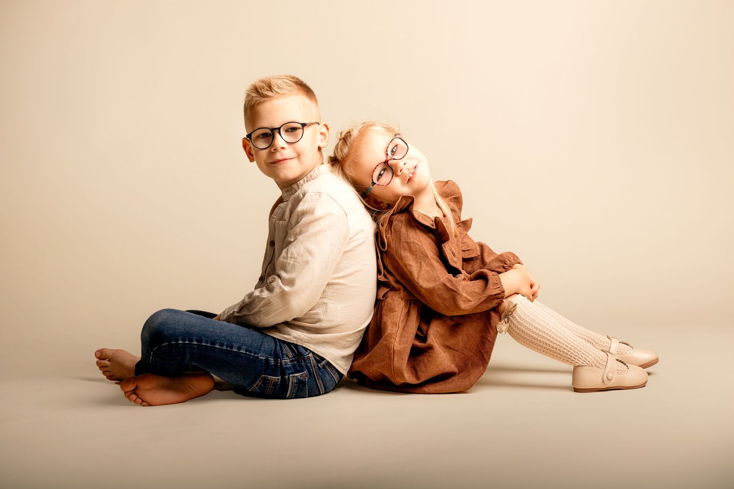 Two young children with glasses sit back-to-back smiling, wearing casual autumn outfits in a studio portrait.
