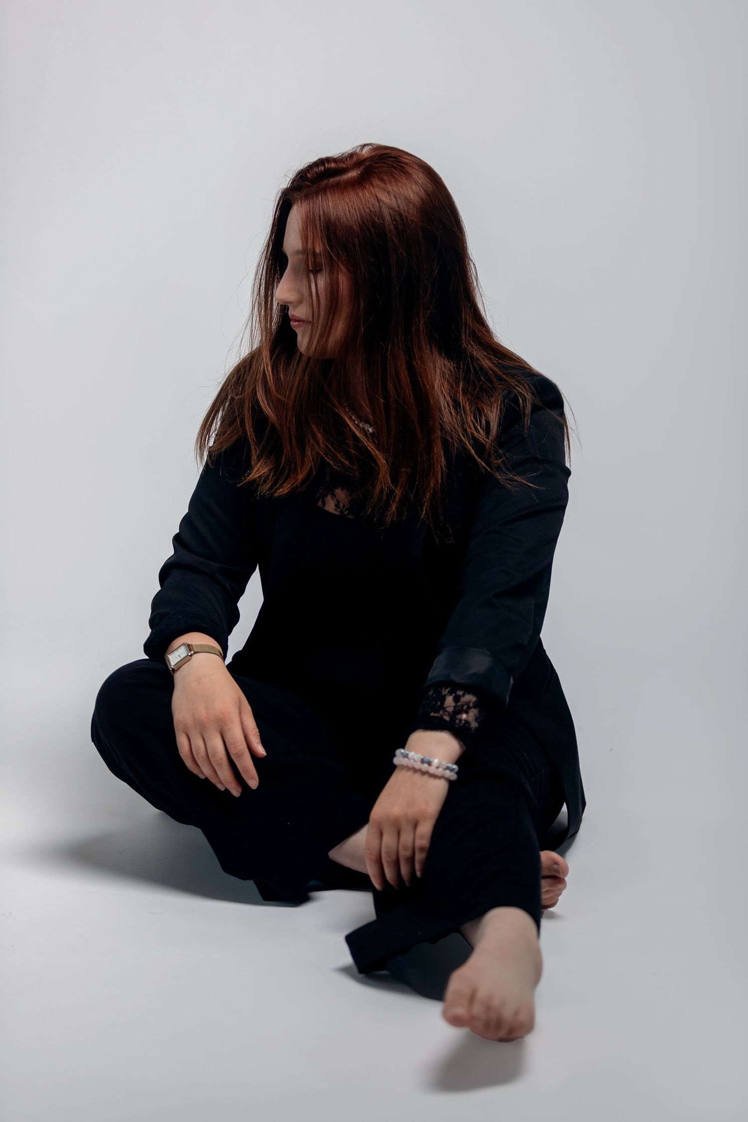 Person in black outfit sits on white background with flowy brown hair and bare feet in moody portrait.