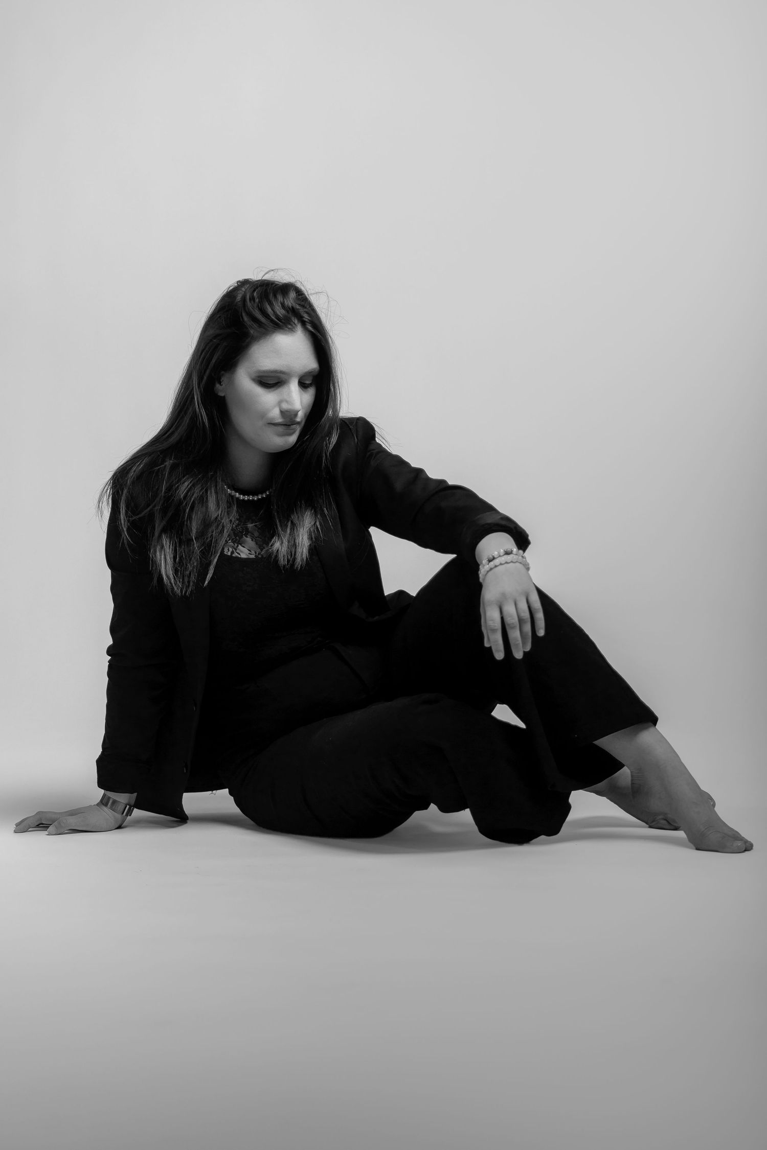 Black and white artistic portrait of a person in dark clothing sitting on floor against white backdrop.