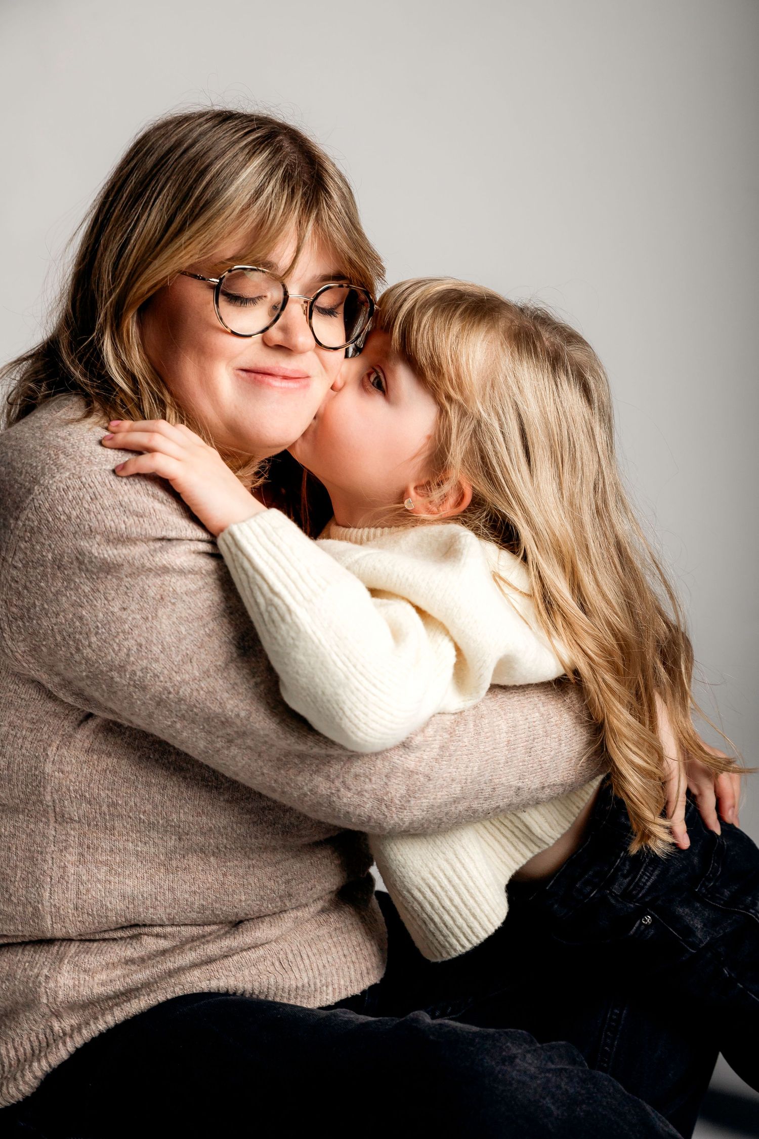 A young girl in a white sweater kisses a smiling woman with glasses wearing a beige sweater in a warm embrace.