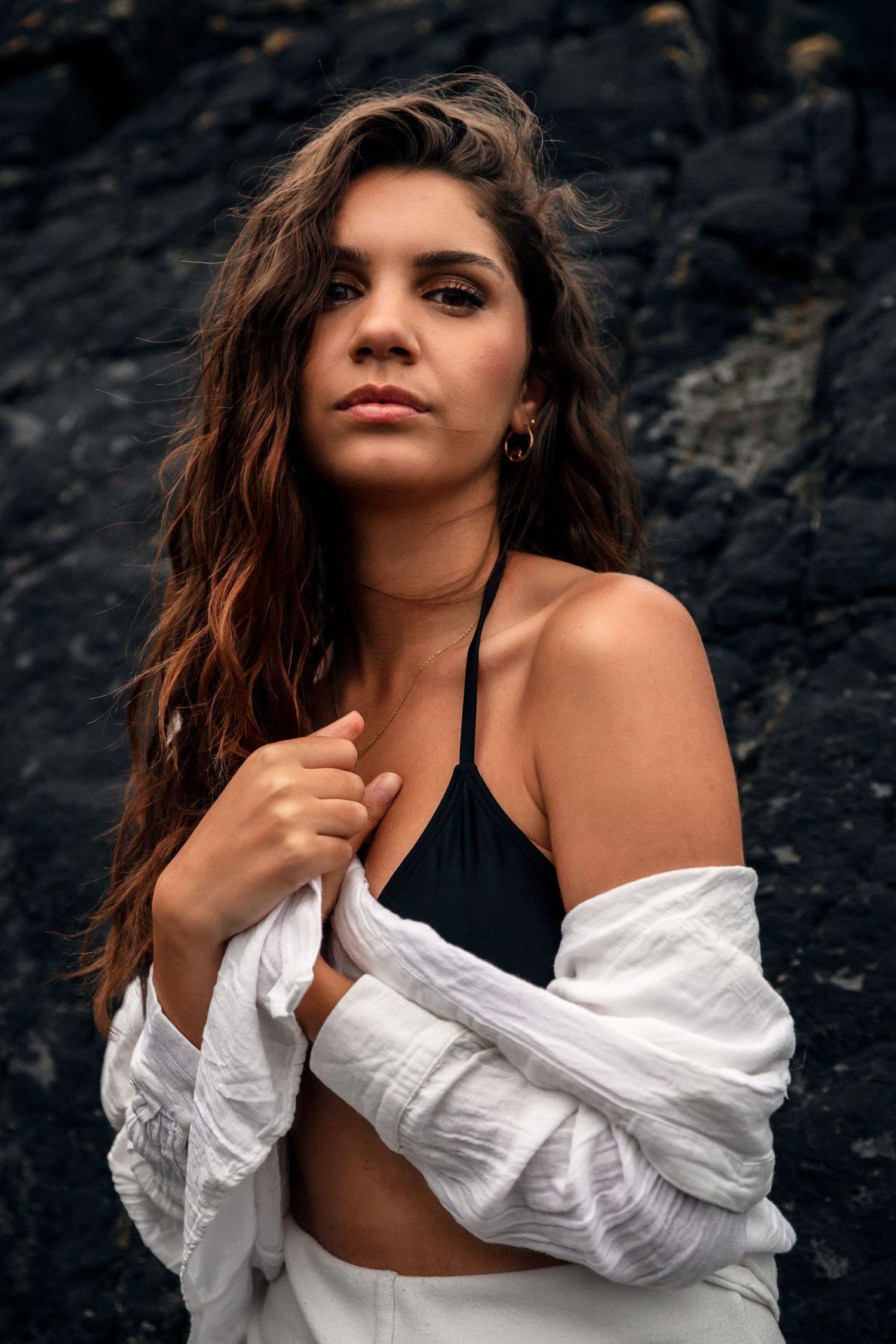 Portrait against dark rocks shows a model in a black top and flowing white shirt with tousled brown hair falling naturally.