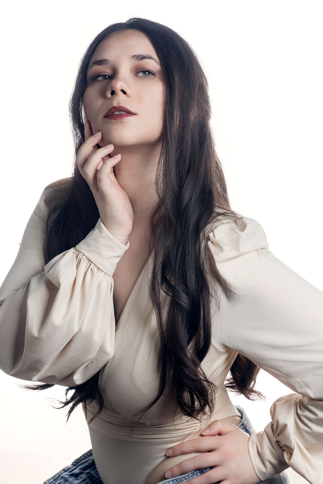 Young woman with long dark hair in cream blouse poses thoughtfully, hand near chin, against white background.
