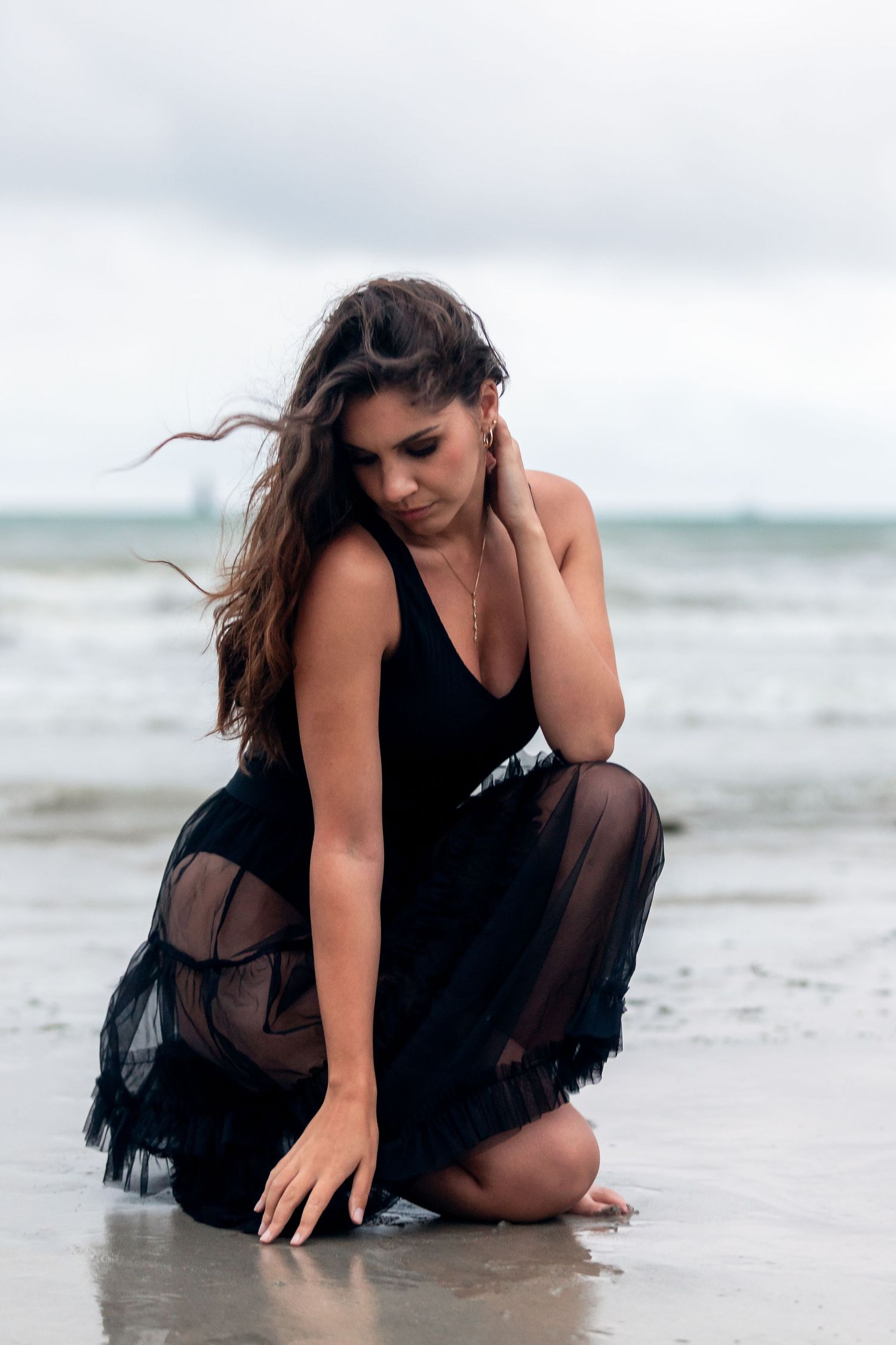 Woman in black dress poses artistically on beach at water's edge in moody photoshoot series.