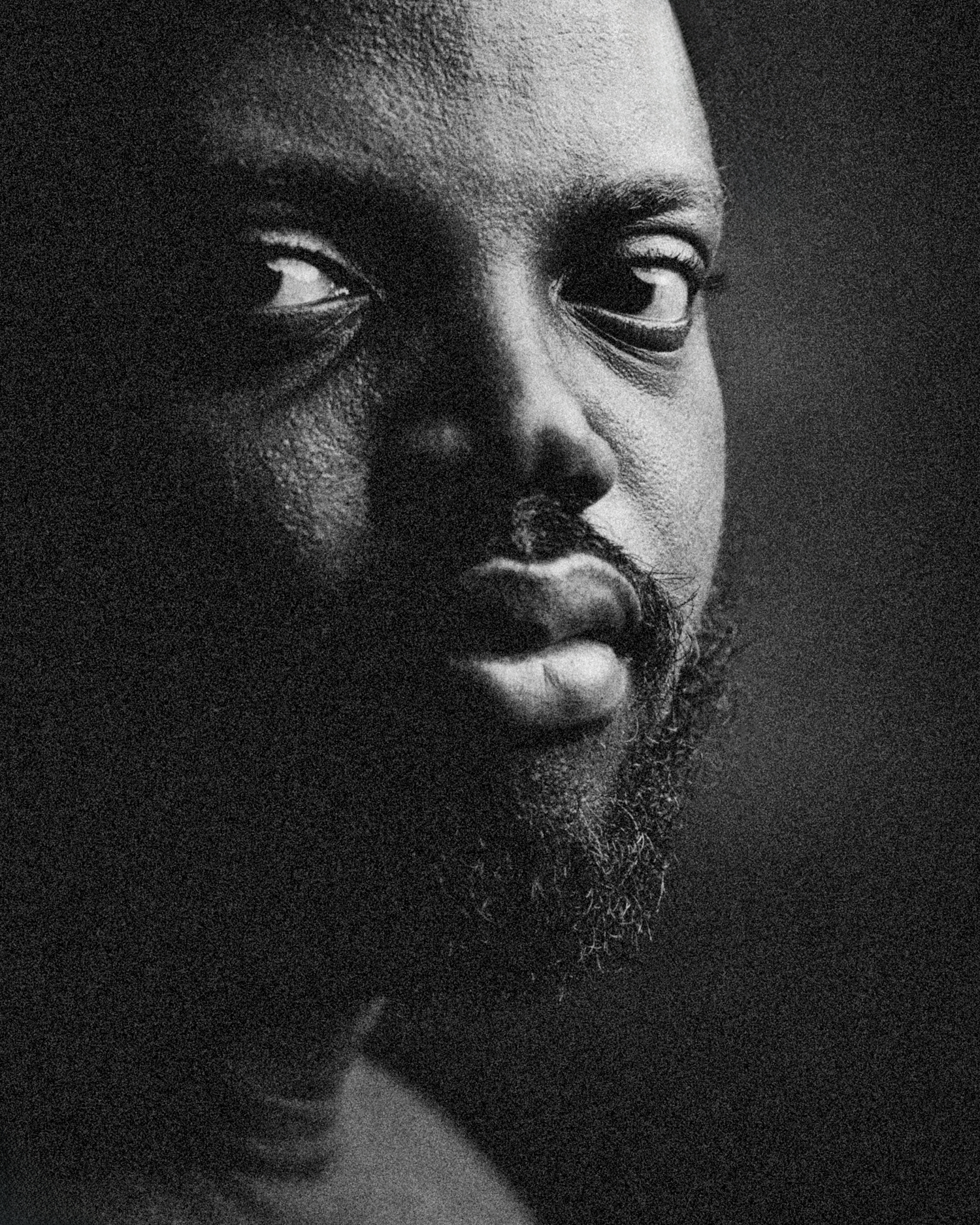 Dramatic black and white close-up portrait of a person with a beard looking to the side against a dark background.