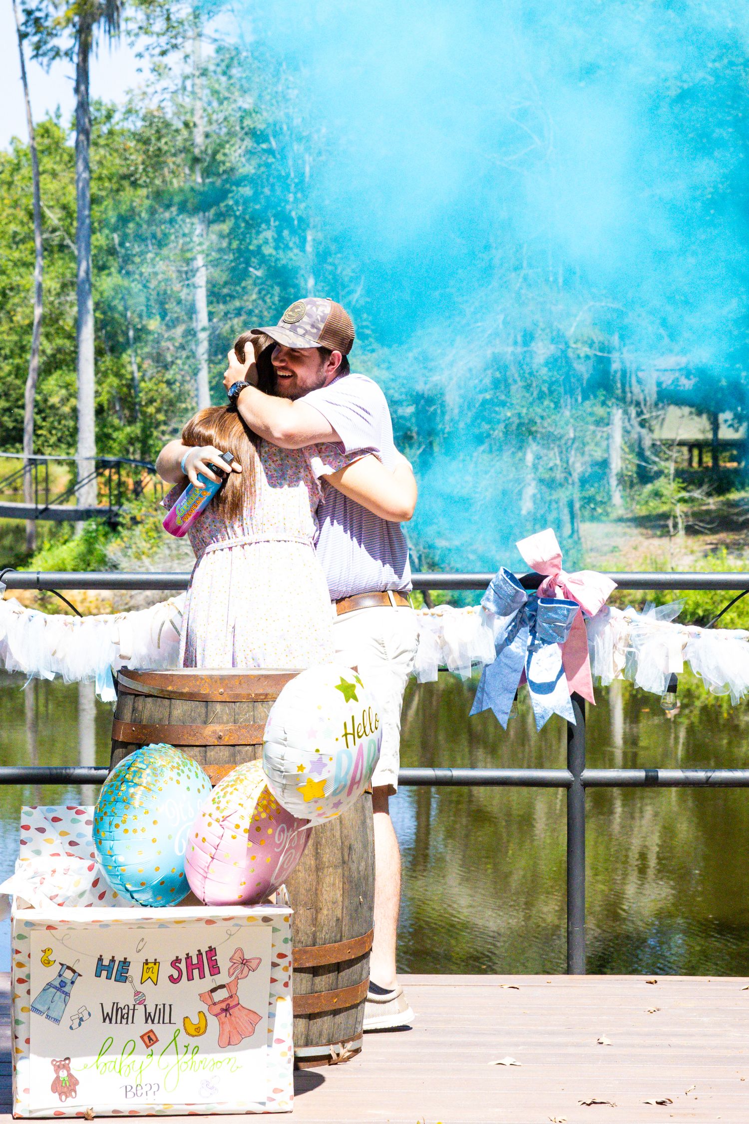 Gender reveal celebration with blue smoke bombs and rustic barrels by a lake creates an exciting outdoor announcement moment.