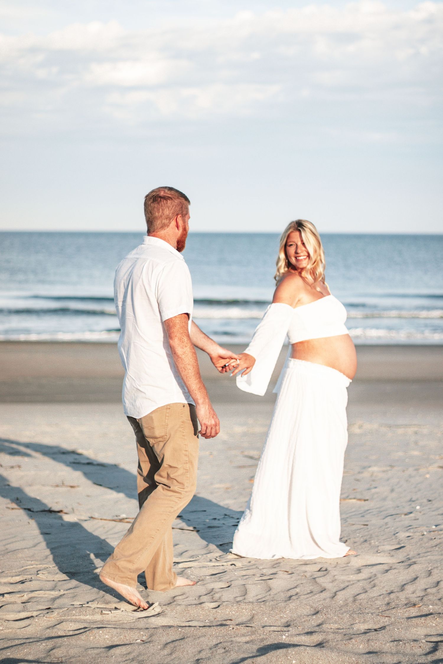 A couple stands together on a sandy beach with ocean waves in the background.