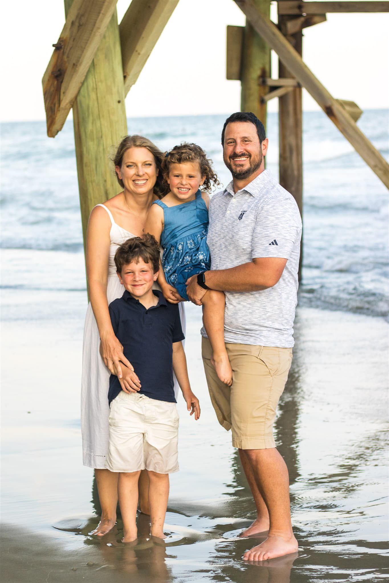 A family stands together in shallow ocean water during a beach photoshoot.