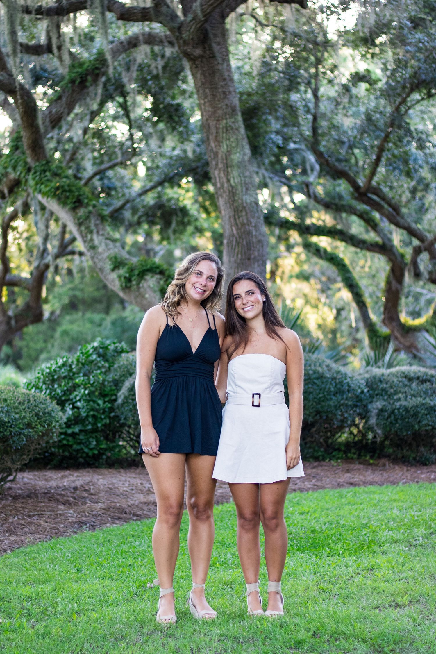 Two figures in coordinated outfits with black and white clothing pose playfully on a grassy lawn surrounded by trees.