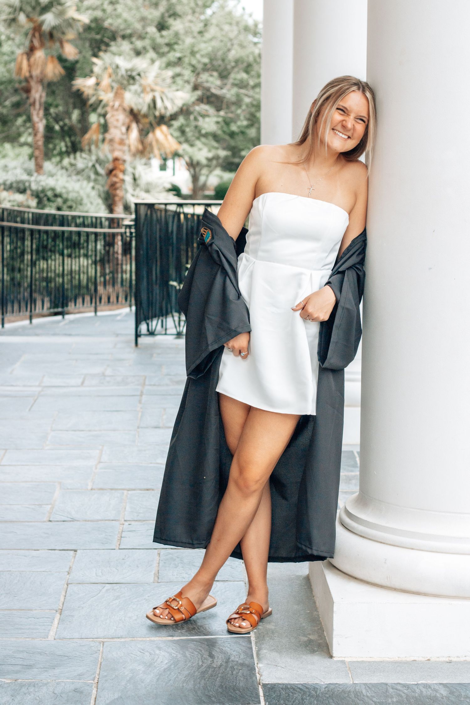 Graduate in white dress and black graduation gown poses against classical column.