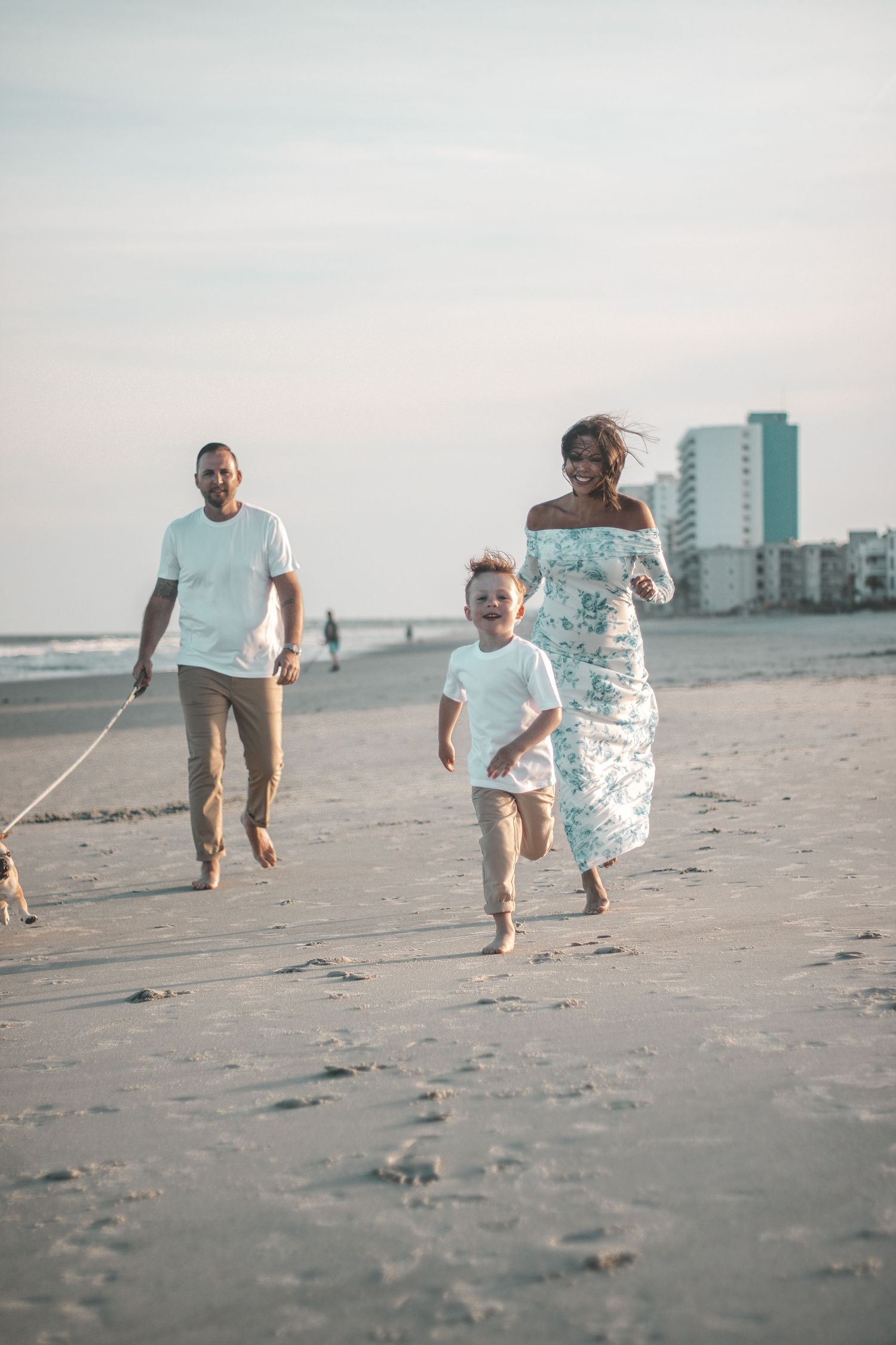 Family running and playing together on a beach at sunset with coastal buildings in background.