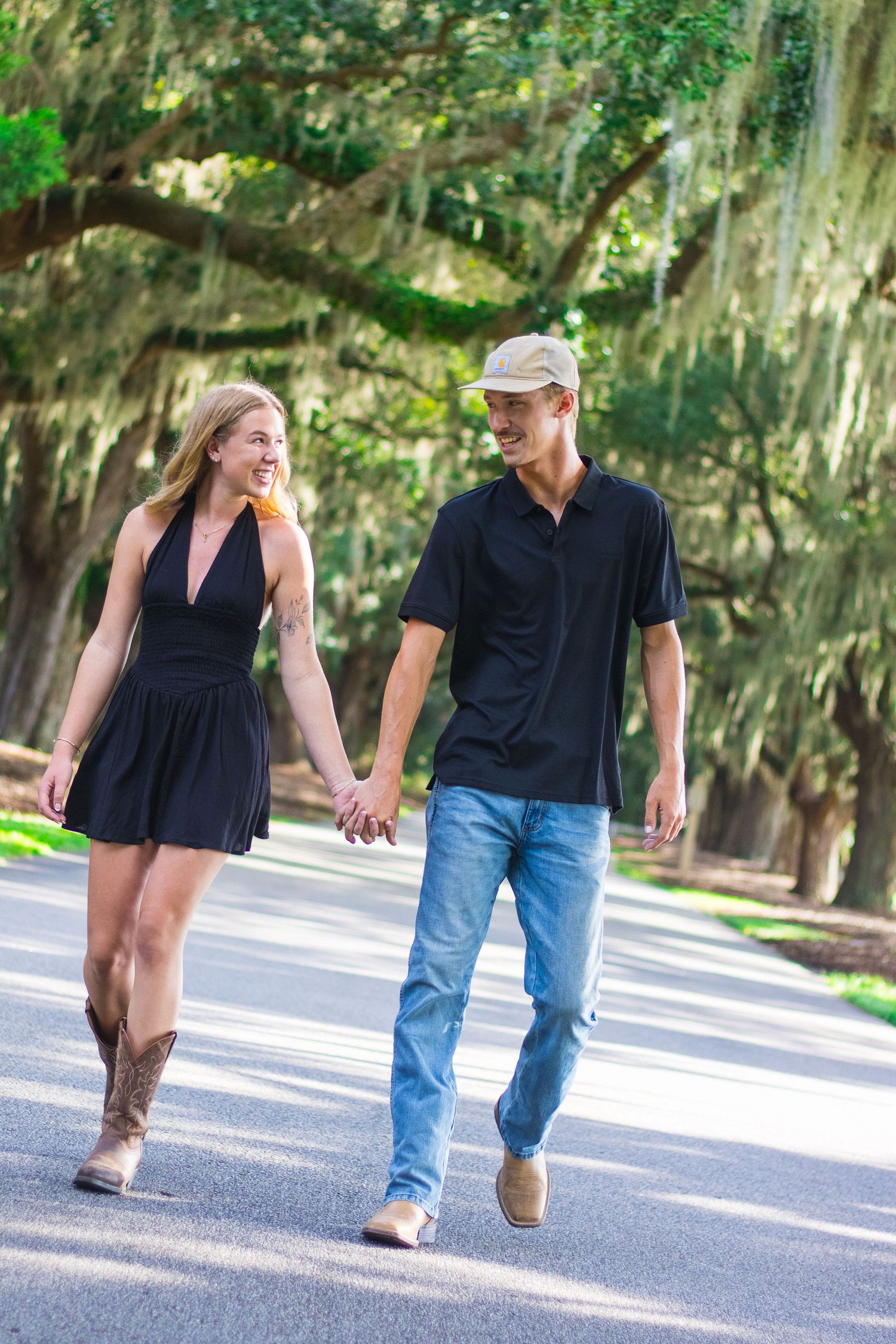 Couple walking hand in hand along a tree-lined path on a sunny day wearing casual attire.